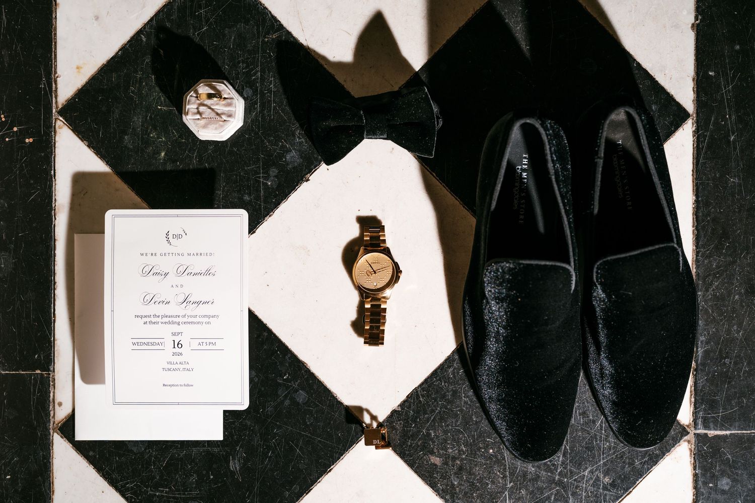Groom's flat lay with black velvet loafers, bow tie, gold watch, ring box and wedding invitation on black and white checkered tile.