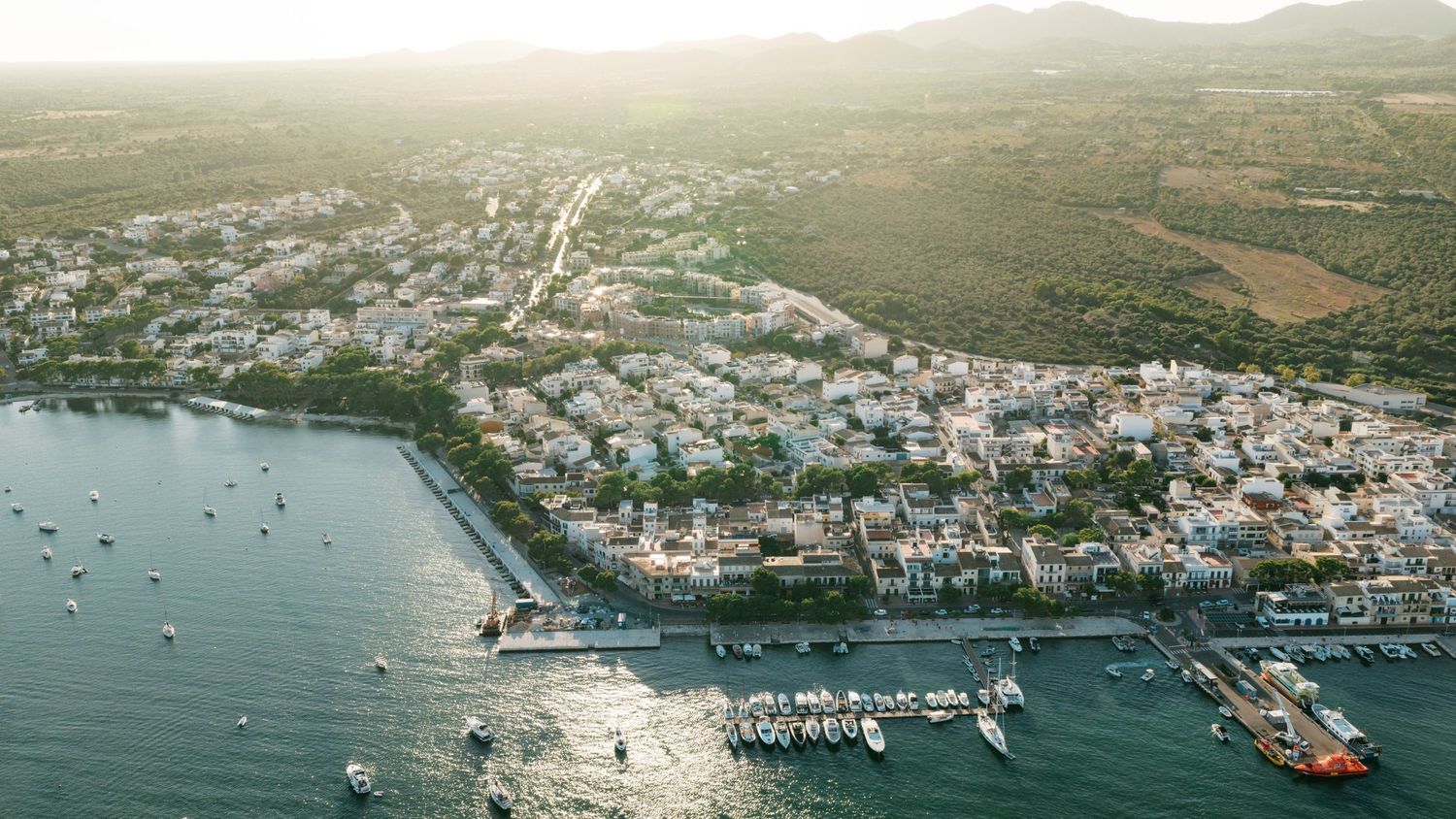 Aerial view of a coastal Mediterranean town with whitewashed buildings, a busy marina filled with boats, and calm blue waters at sunset.