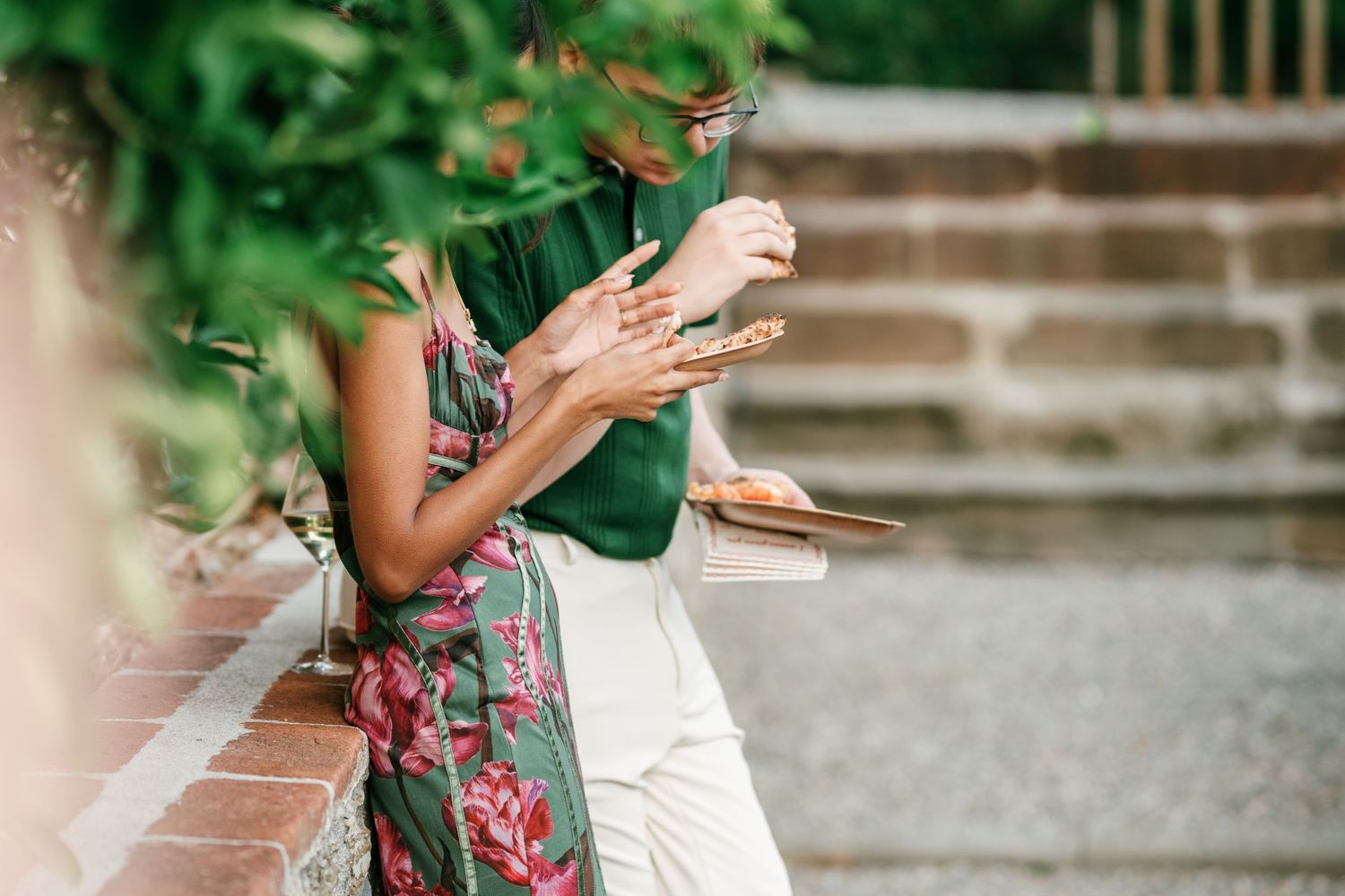 Two women in floral and green summer outfits stand close together outdoors, holding phones beside brick steps and leafy greenery.