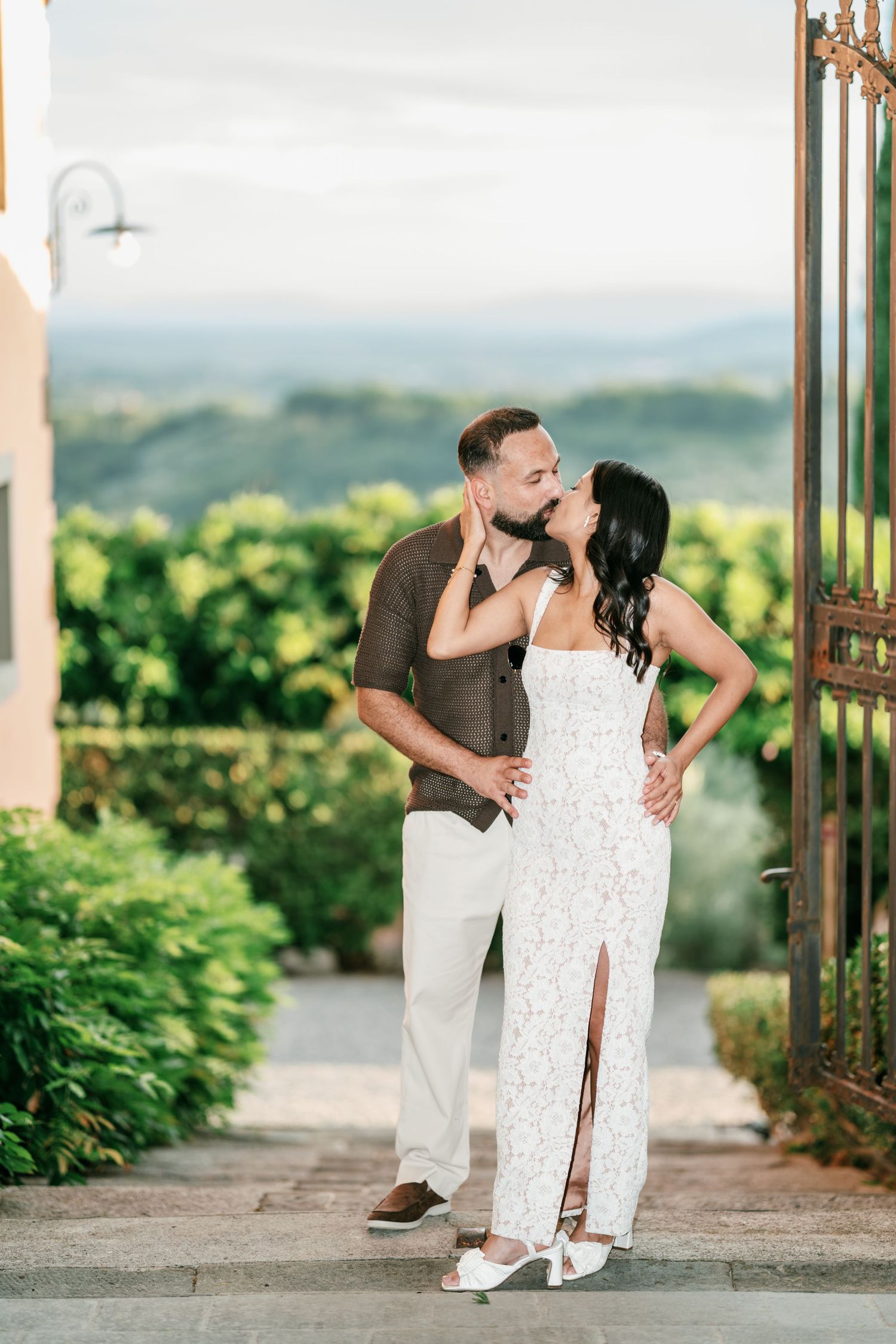 Romantic couple kissing on stone steps beside an iron gate, with lush green hedges and rolling hills in the background.