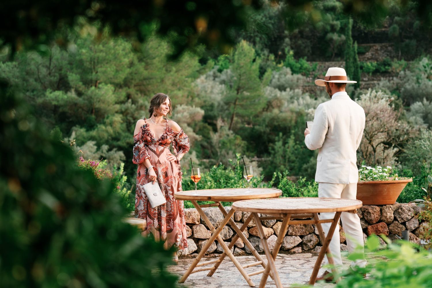 Woman in a floral dress and man in a white linen outfit and straw hat stand beside rustic wooden tables in a lush garden.