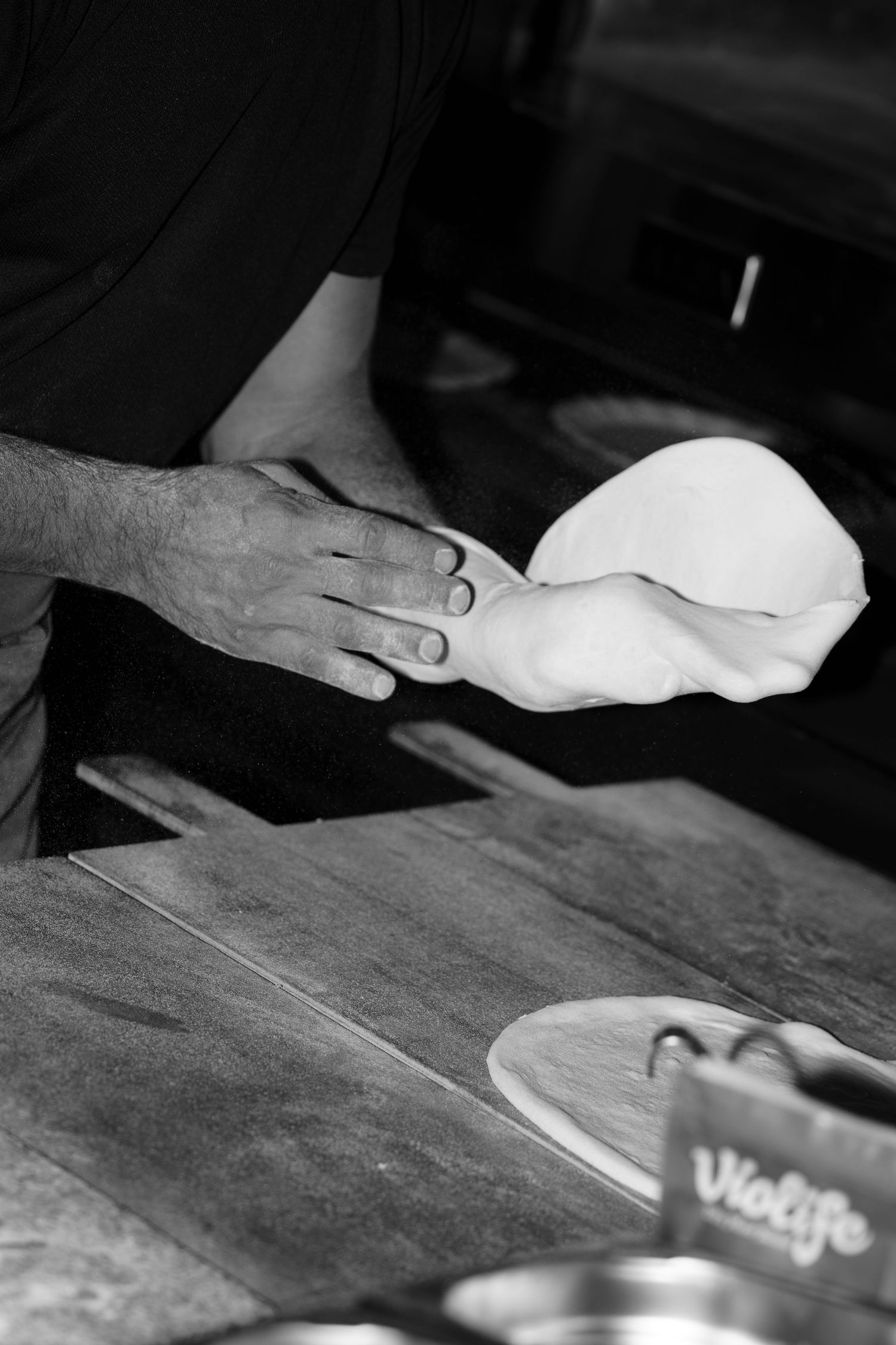 Black and white photo of a baker's hands stretching fresh pizza dough above a rustic wooden counter in a restaurant kitchen.