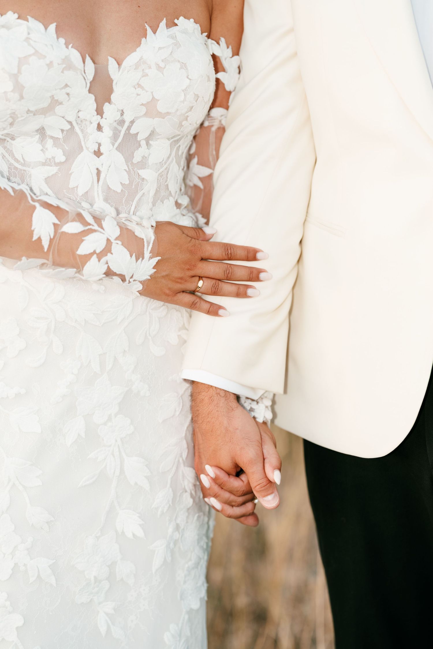 Bride and groom holding hands, showcasing wedding rings, bride in lace floral gown, groom in white tuxedo jacket.