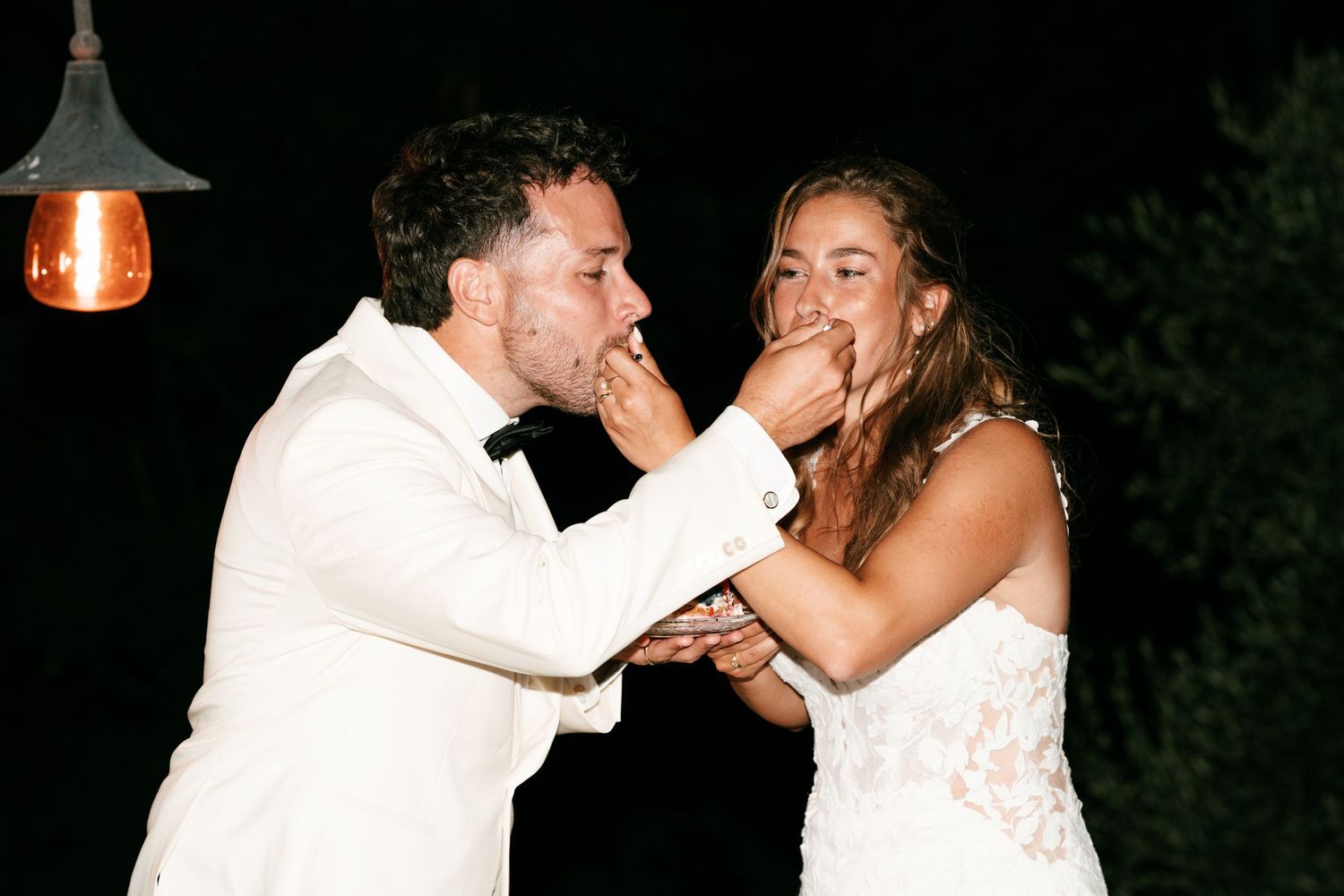 Bride and groom feeding each other wedding cake at night outdoor reception, laughing joyfully.