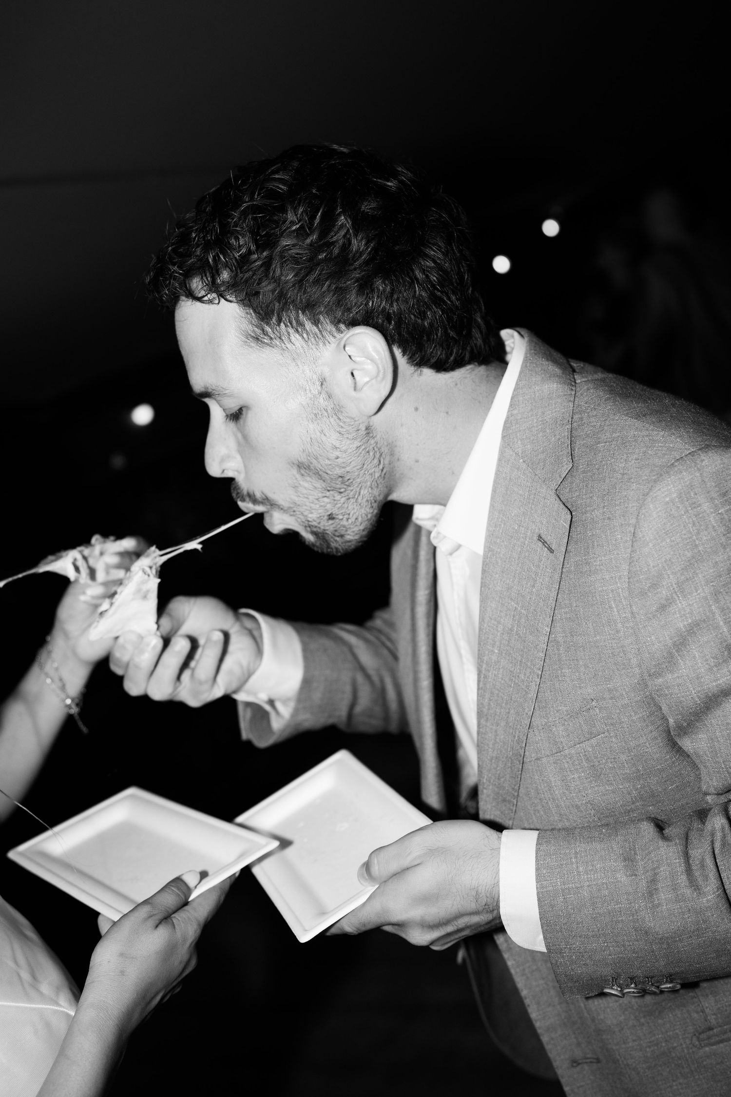 Groom eating wedding cake in black and white photo at reception celebration.