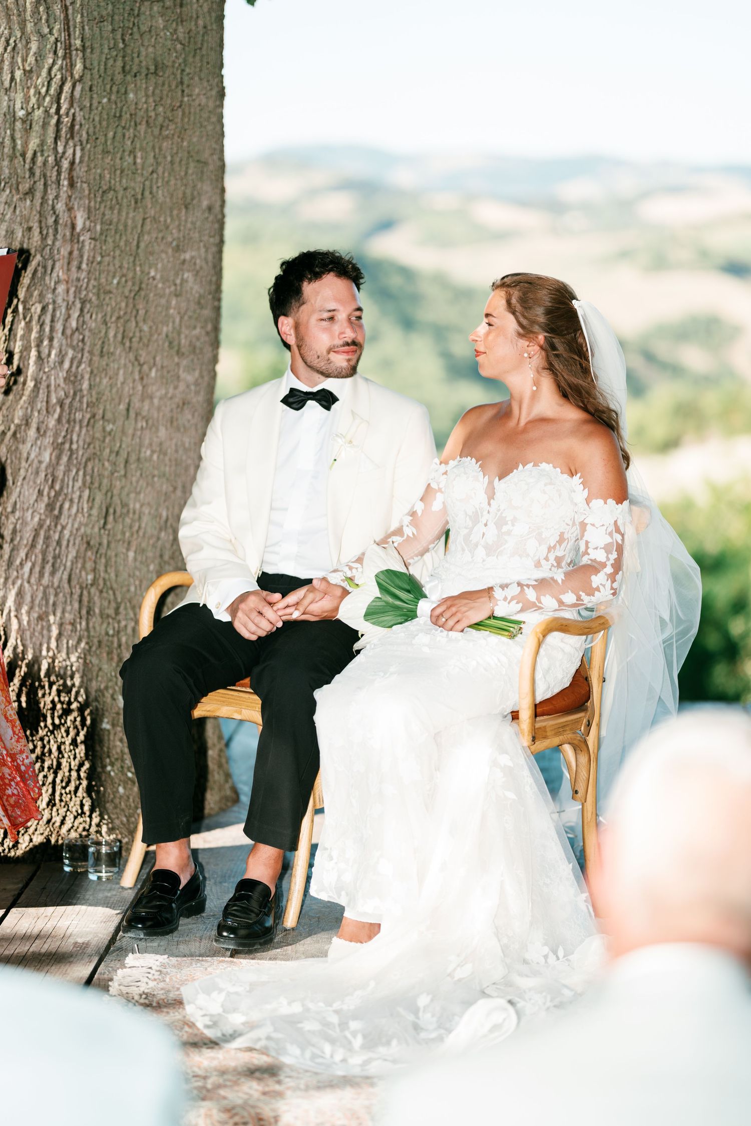 Bride and groom sit together during outdoor wedding ceremony beneath large tree with scenic valley views behind them.