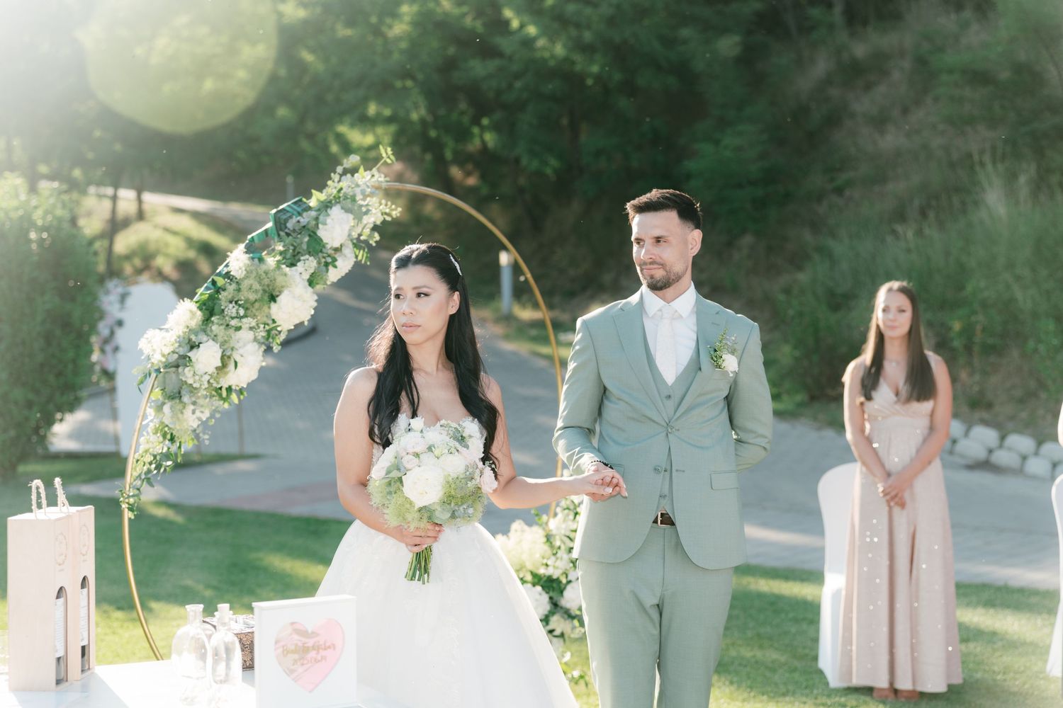 Bride and groom stand at floral hoop altar during outdoor wedding ceremony on sunny green lawn. Italy wedding photographer and filmmaker