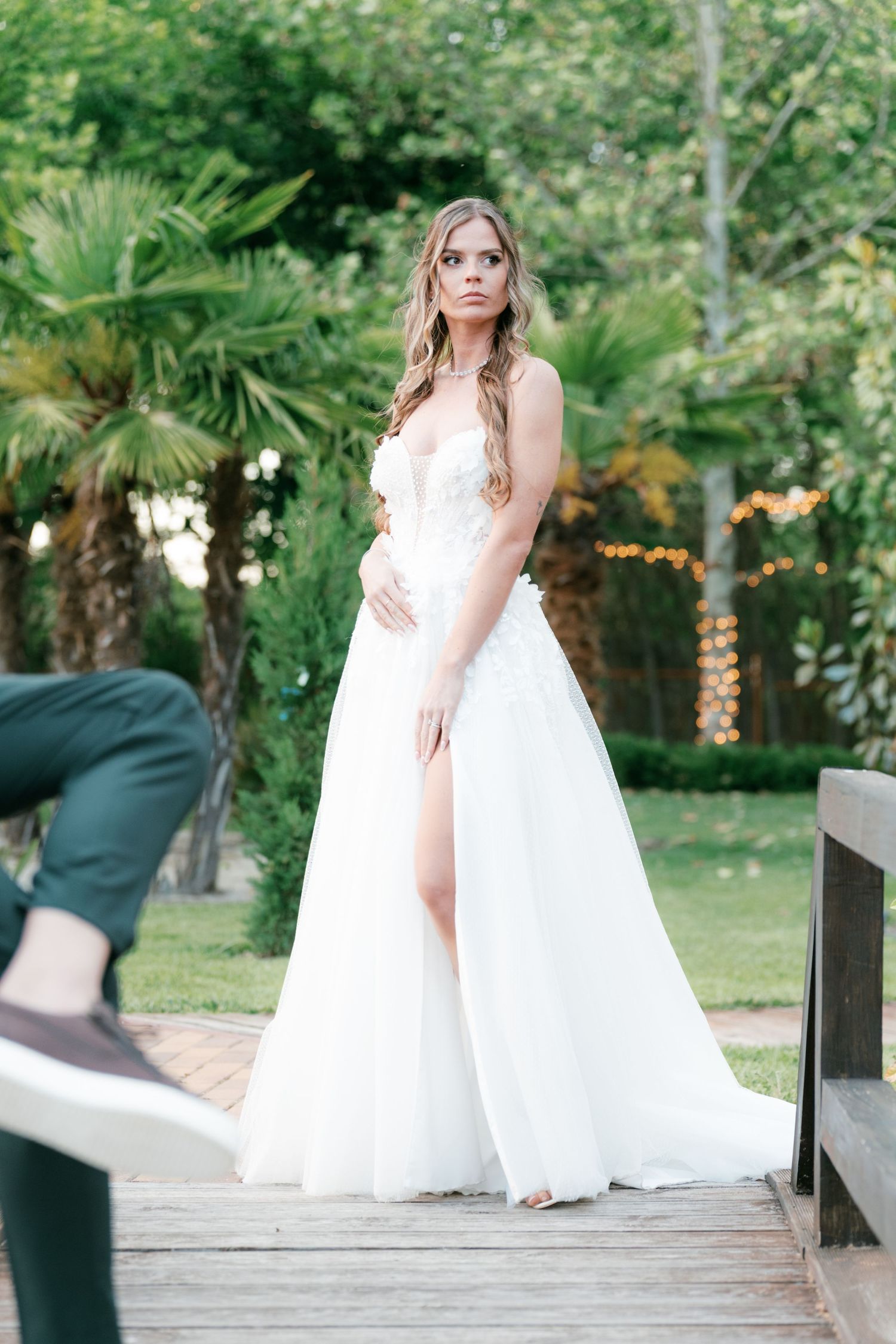 Bride in flowing white wedding gown with thigh slit poses on wooden deck in tropical garden. Mallorca wedding photographer and filmmaker.