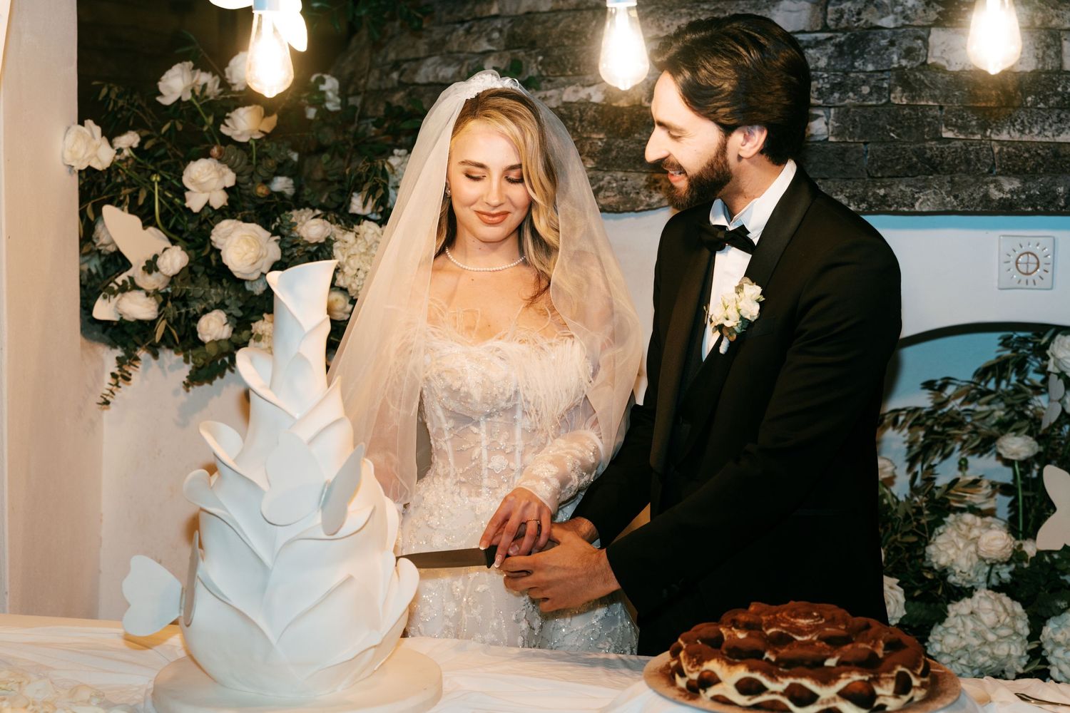 Bride and groom cutting a white tiered wedding cake surrounded by floral arrangements. Puglia wedding photographer, film