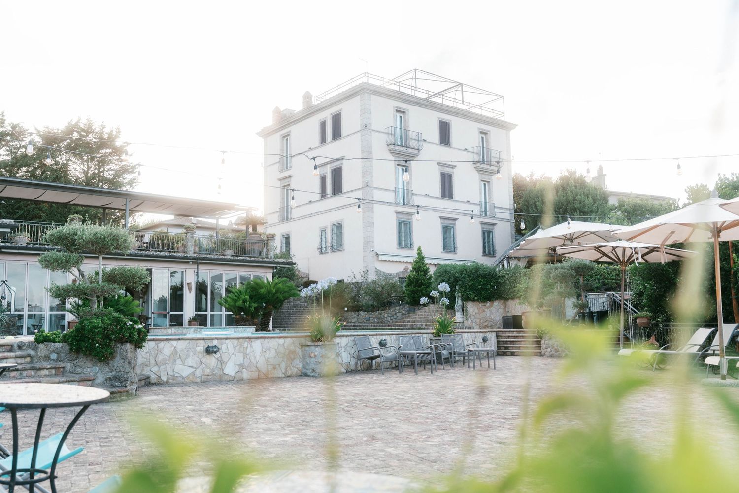 Elegant white Italian villa with outdoor terrace, umbrellas, and lush greenery in soft evening light. Puglia wedding photographer, film