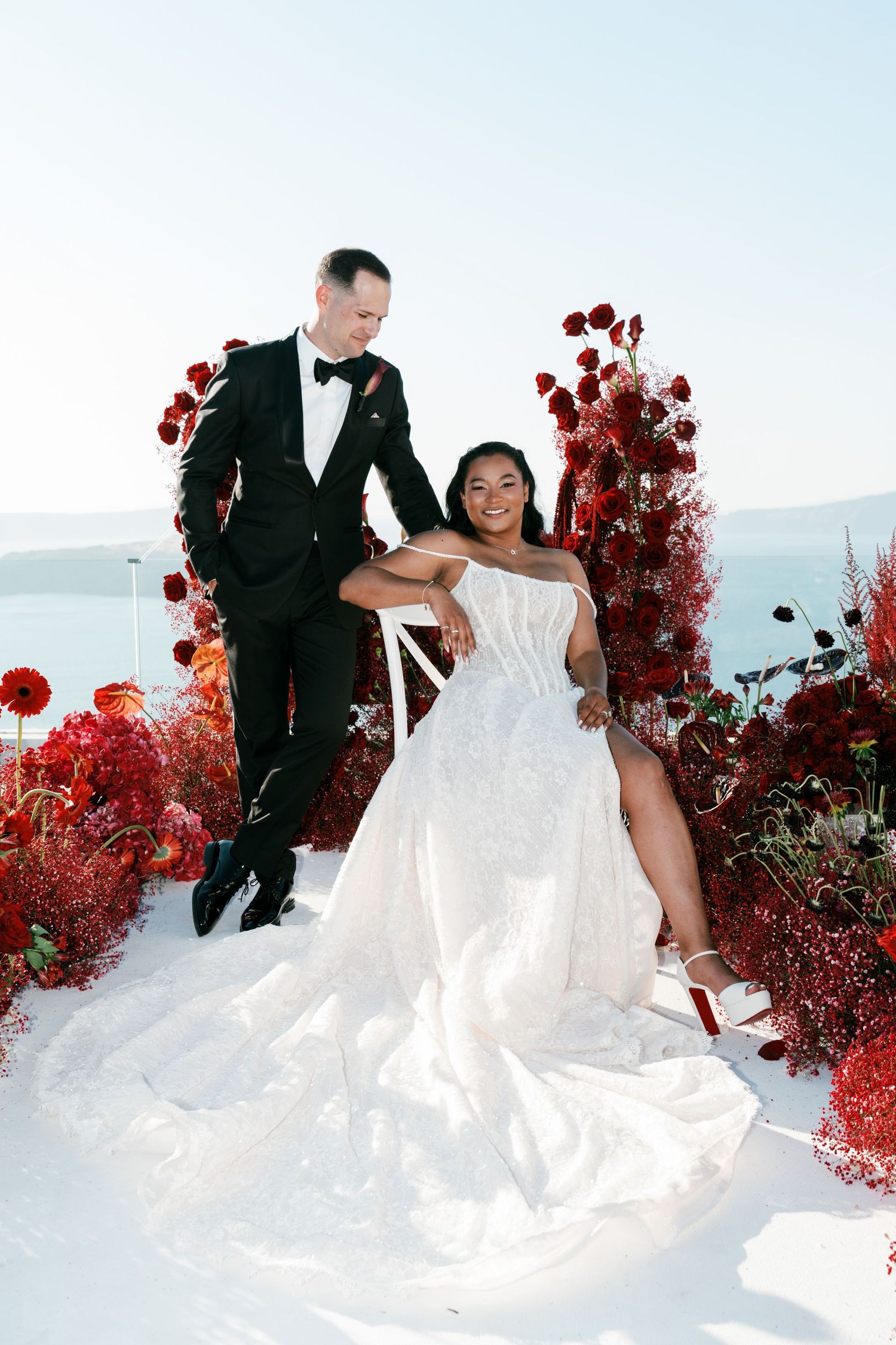Bride in white strapless gown and groom in black tuxedo pose among red floral arrangements with ocean view backdrop.