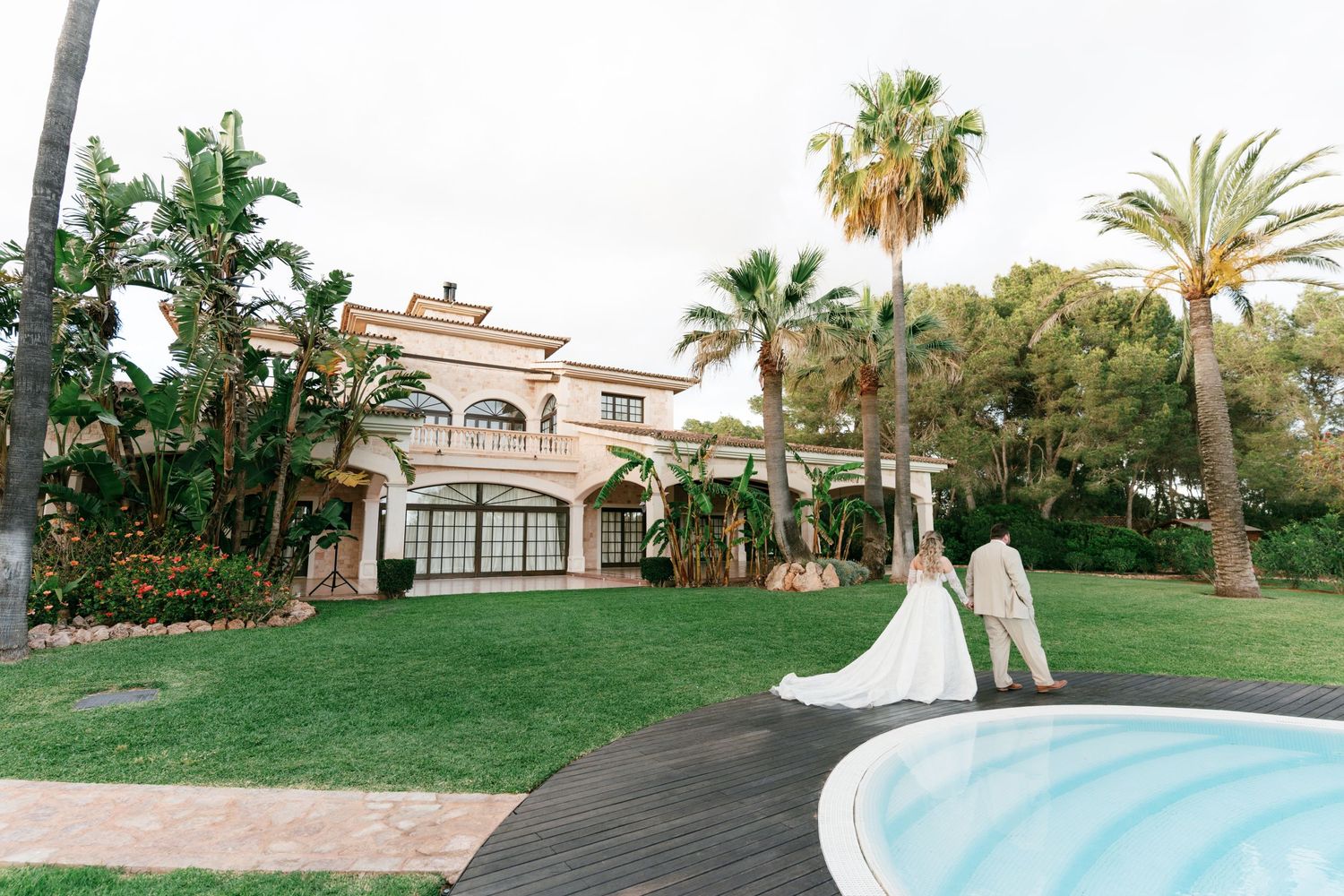 Bride and groom stand by a pool at a luxurious Mediterranean villa surrounded by palm trees and lush greenery in Mallorca.