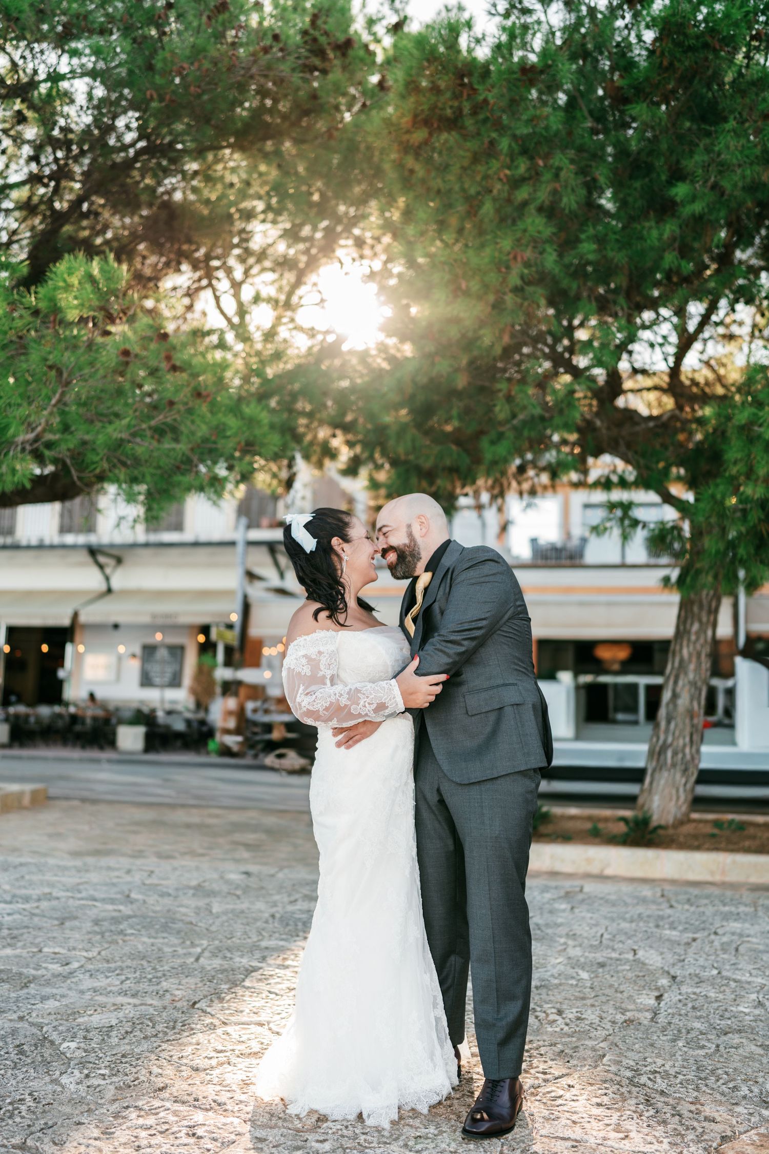 Bride in a lace wedding gown and groom in a gray suit embrace under pine trees as the sun sets over a seaside promenade.