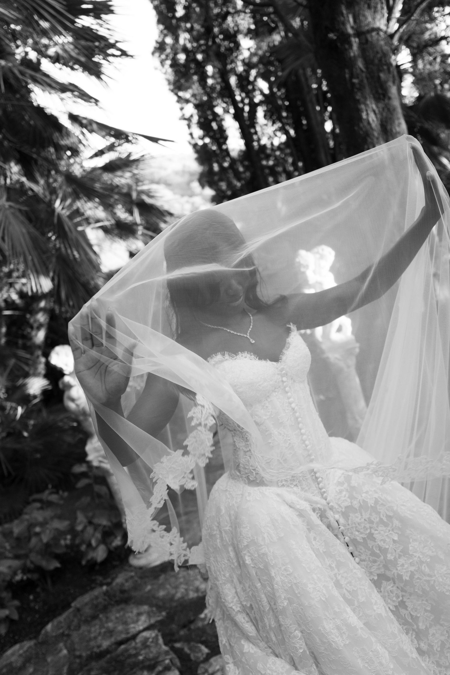 Black and white photo of a bride in a lace wedding gown lifting her sheer veil overhead amid lush garden trees and rocks.