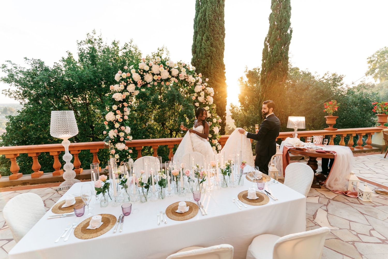 Elegant outdoor wedding table with gold chargers and glass lamps beside a floral arch, as a couple stands on a terrace at sunset.