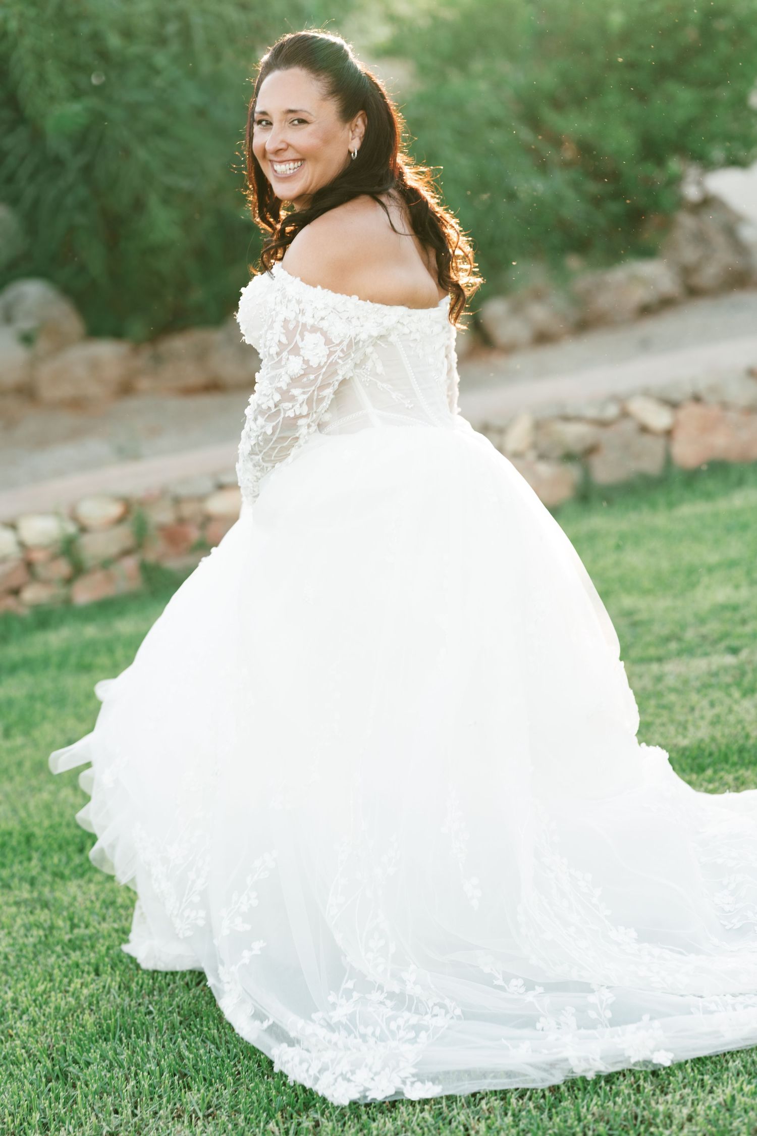 Smiling bride in an off-the-shoulder lace wedding dress with a long tulle train looks back over her shoulder on a sunny lawn.