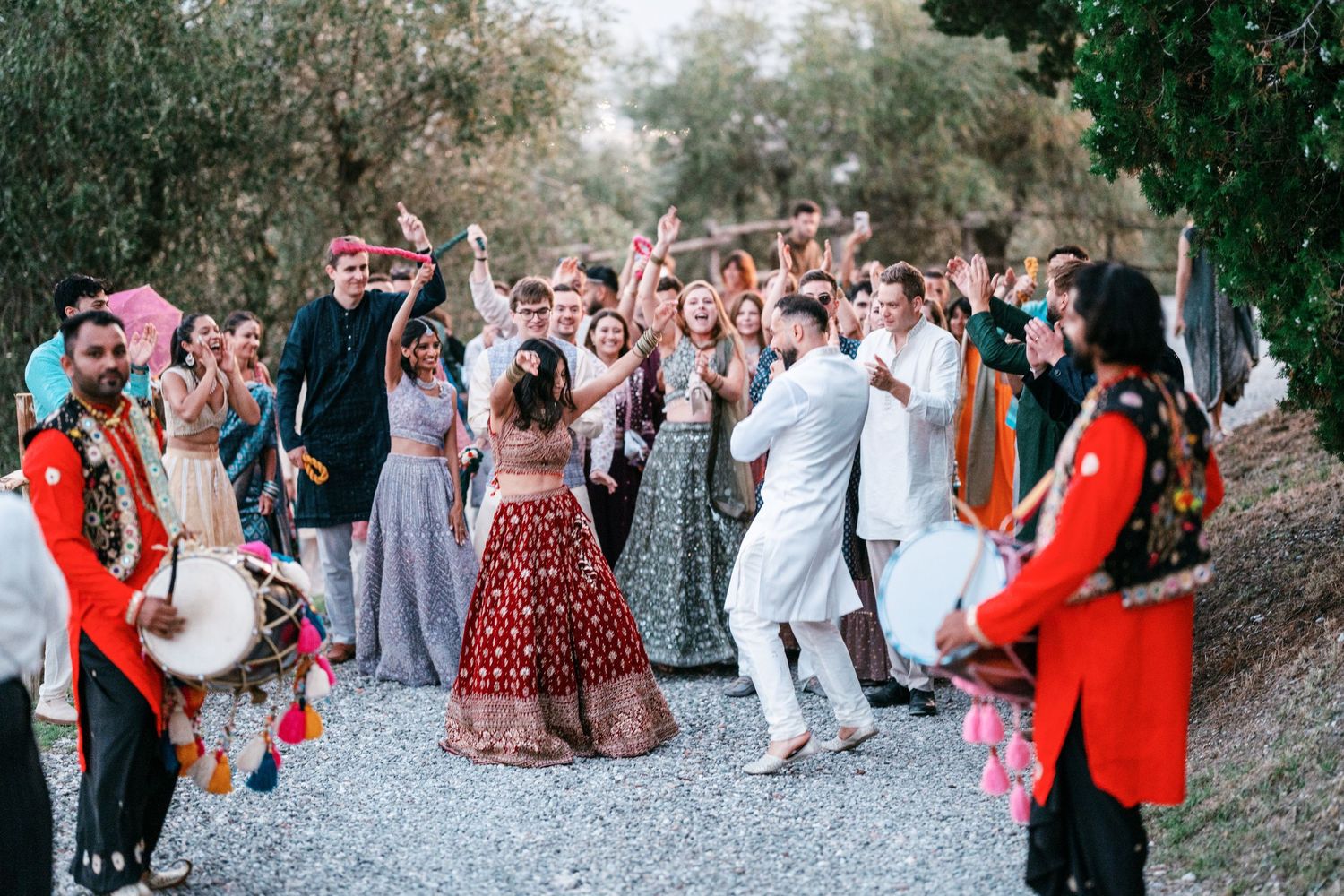 Wedding guests dance and cheer on a gravel path as dhol drummers in red outfits lead a lively Indian baraat procession outdoors.