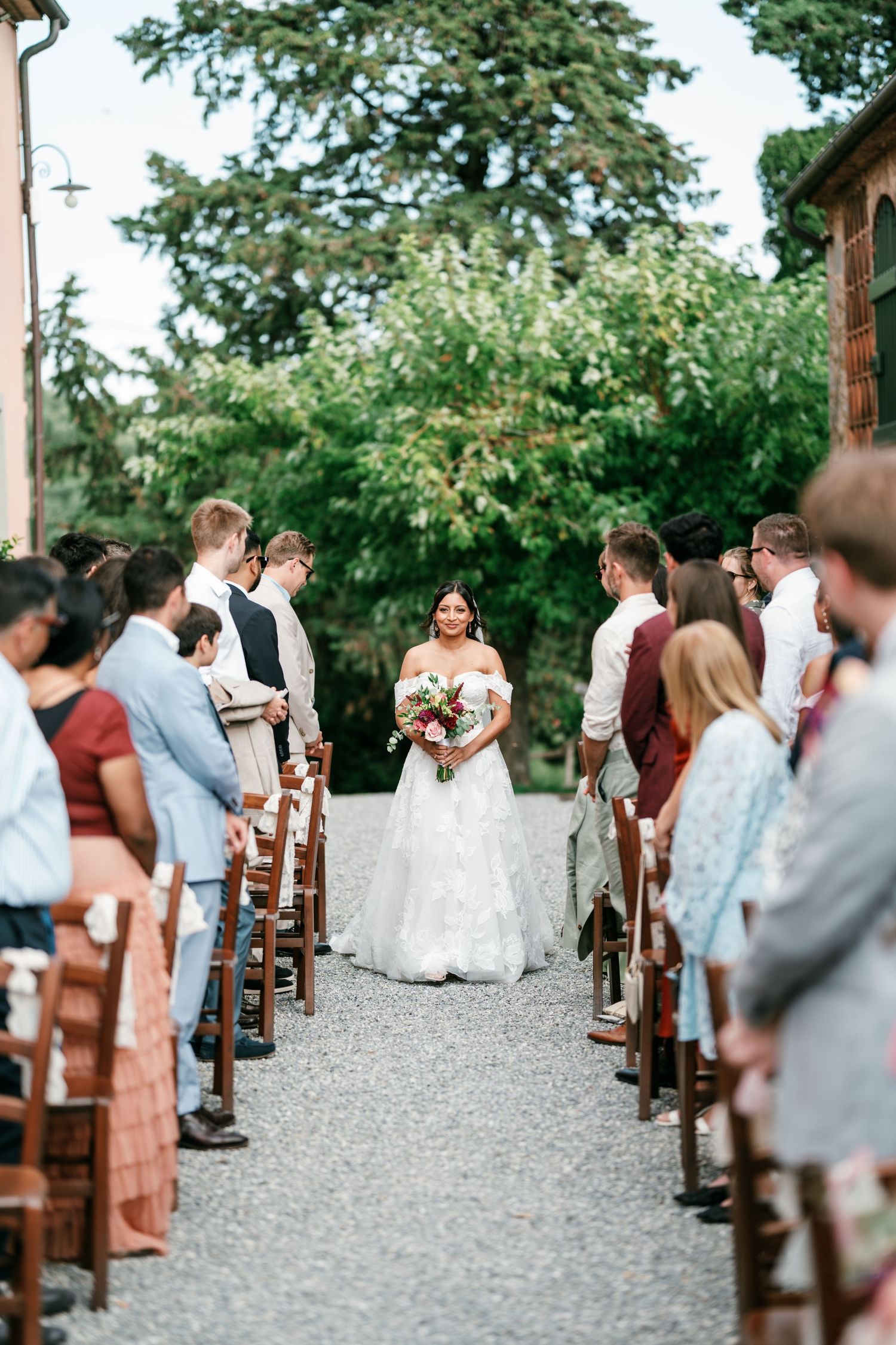 Bride in an off-shoulder wedding gown carrying a bouquet walks down a gravel aisle between seated guests at an outdoor garden ceremony.