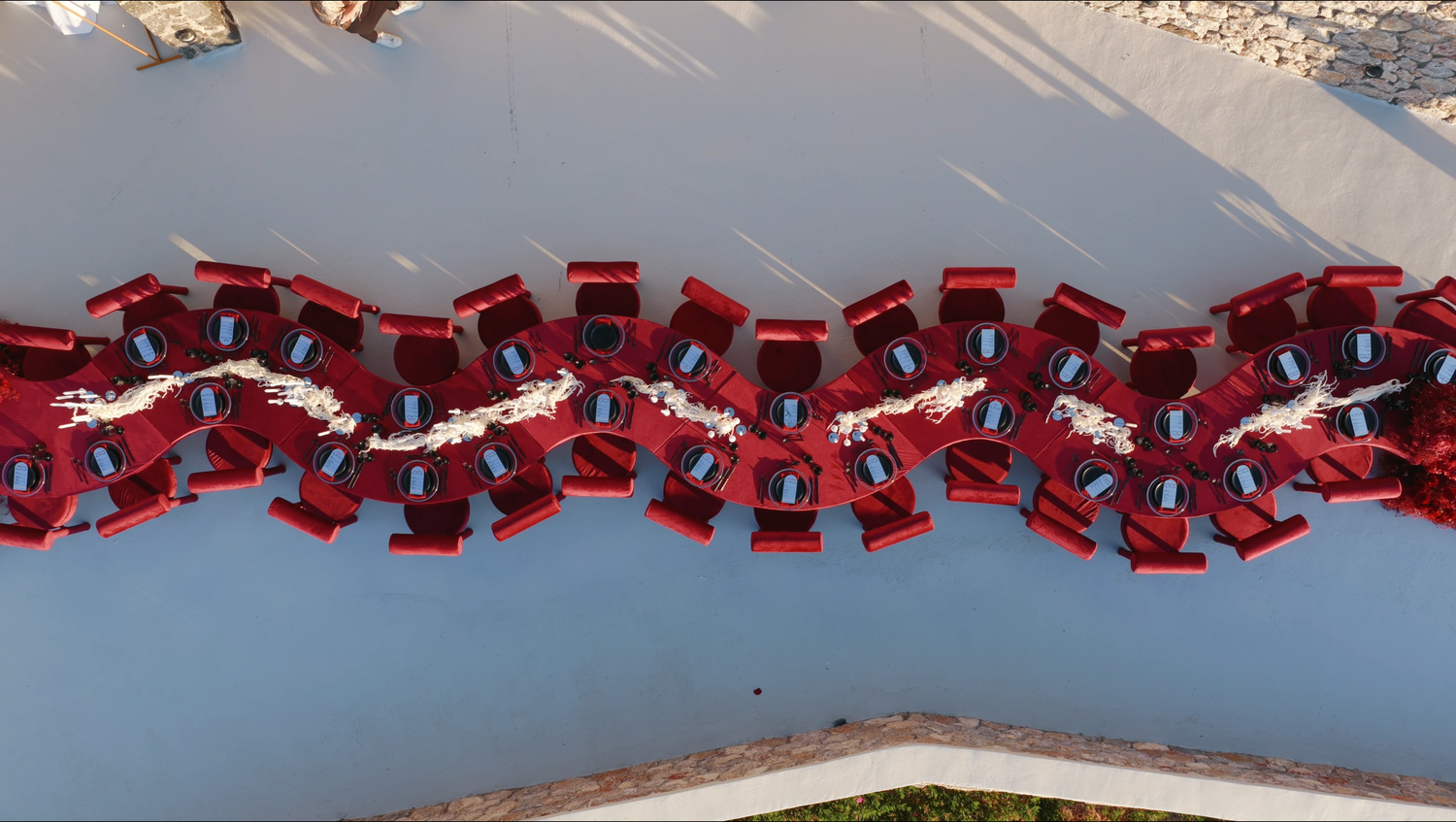 Aerial view of a long outdoor wedding reception table with red chairs, white floral runners, and blue place settings on a pale terrace.