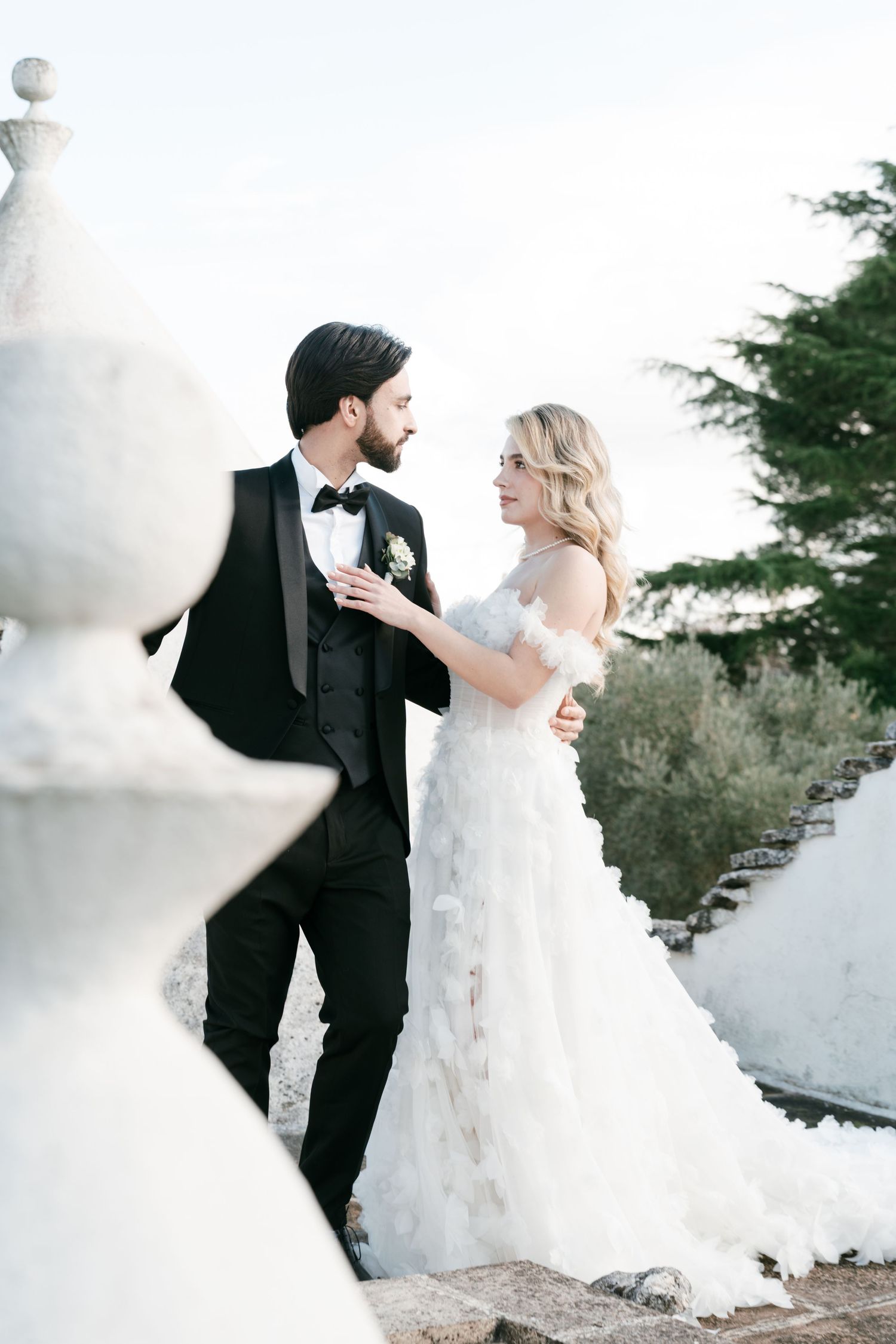 Bride in a ruffled off-shoulder gown and groom in black tuxedo sharing a romantic moment on a stone terrace.Puglia wedding photographer,