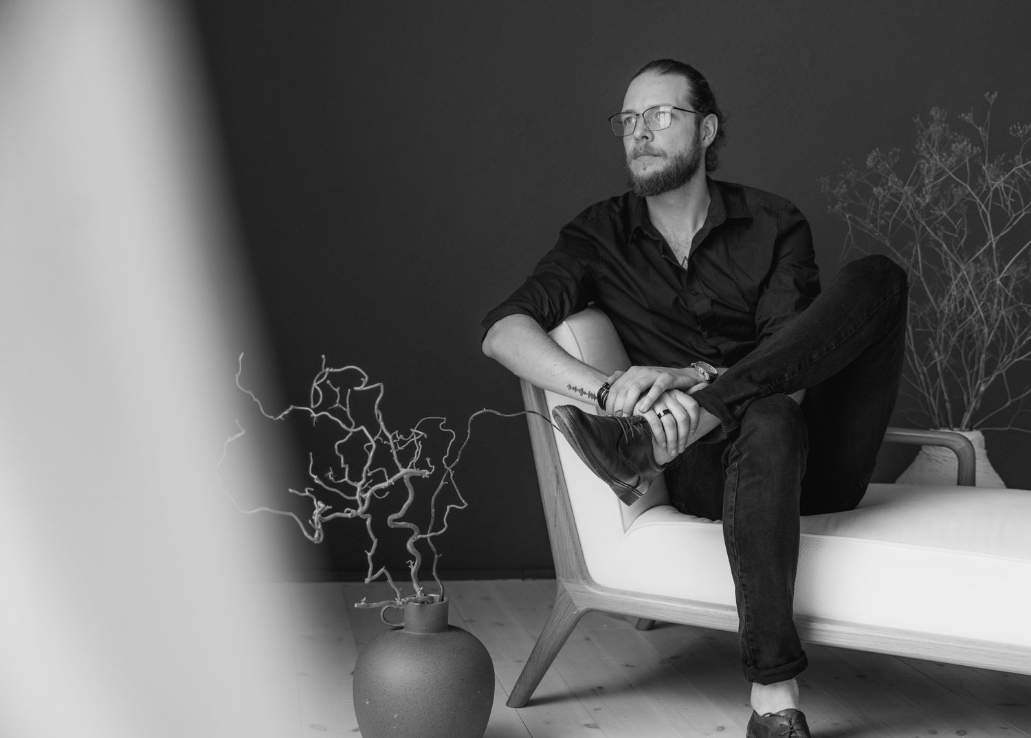 Black and white portrait of a bearded man in black shirt sitting thoughtfully on a modern white chaise lounge. Mallorca wedding photographer