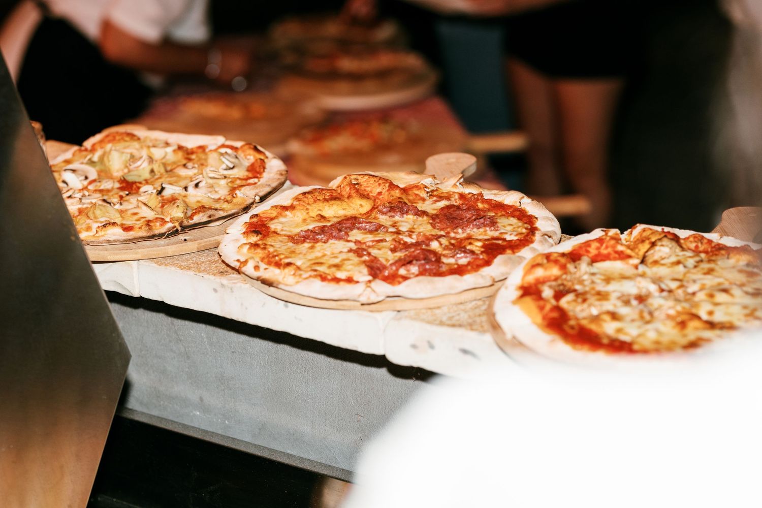 Three freshly baked pizzas with various toppings displayed on a wooden counter in a busy pizzeria.