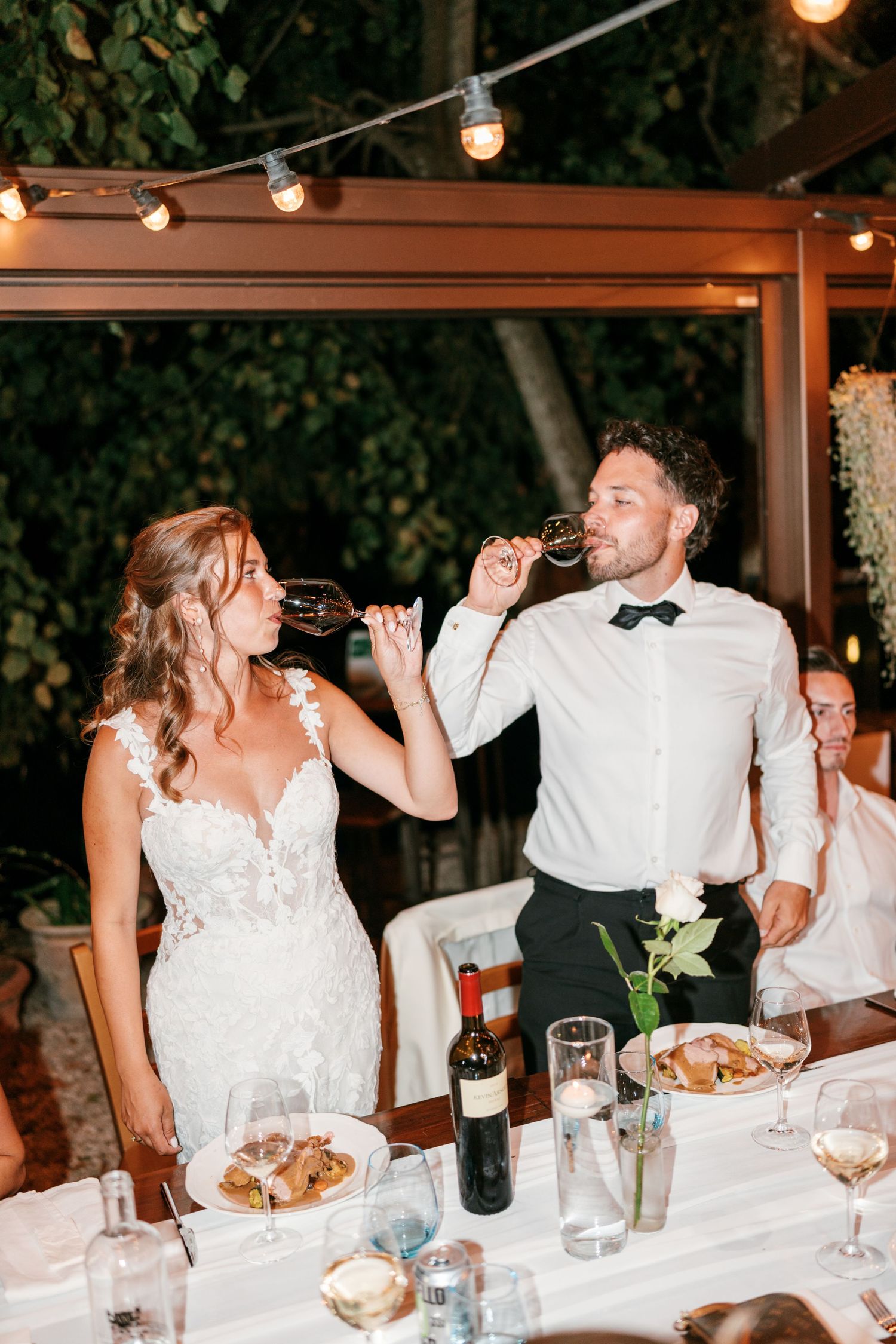 Bride and groom toast with wine glasses at outdoor evening wedding reception under string lights.