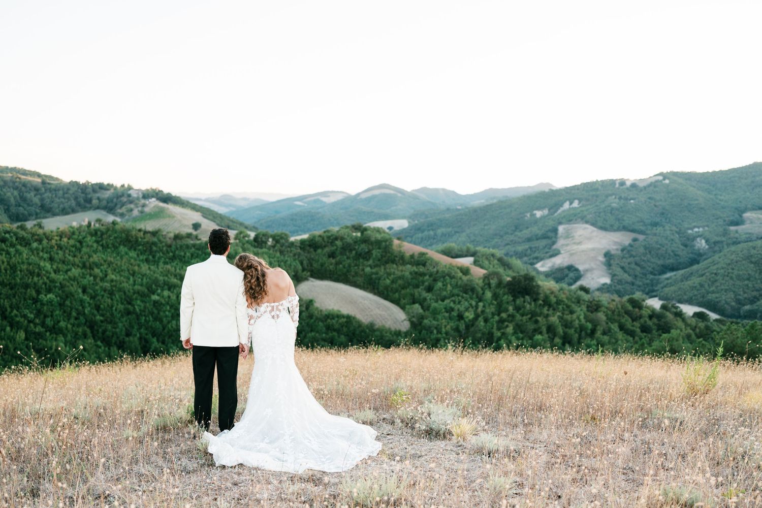 A bride and groom stand together overlooking rolling green hills and golden meadows in a romantic outdoor wedding photo.