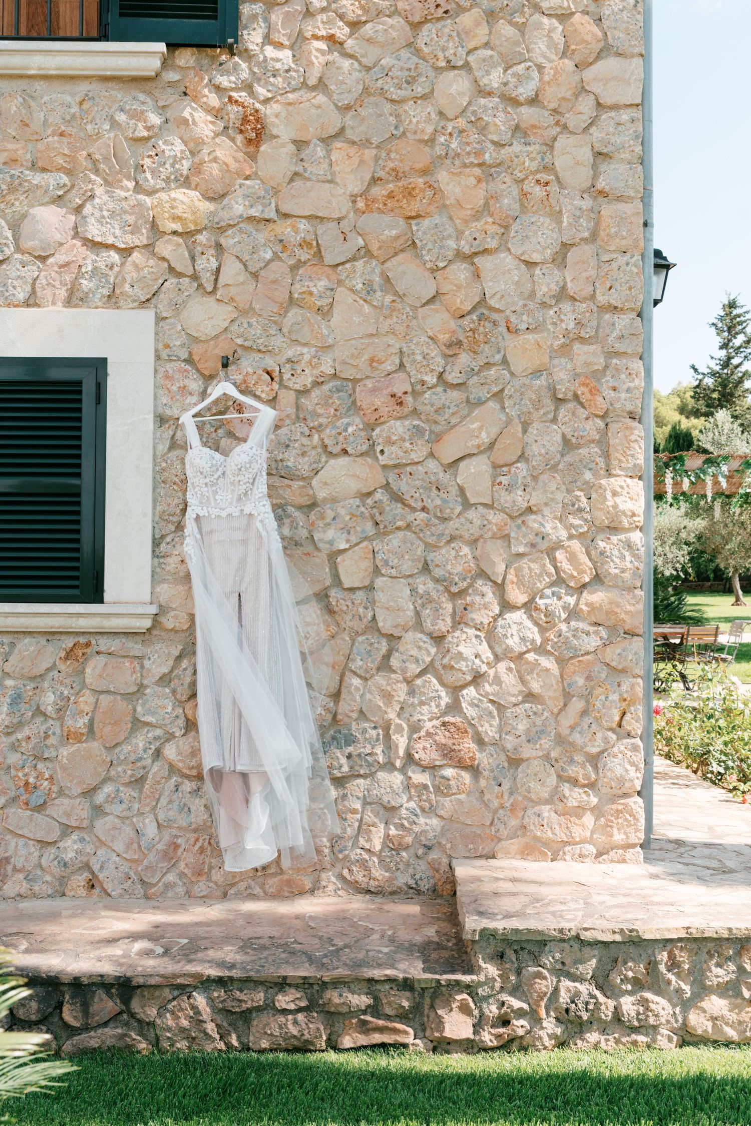 Elegant white lace wedding dress hanging on a rustic stone building exterior wall. Mallorca wedding photographer, filmmaker