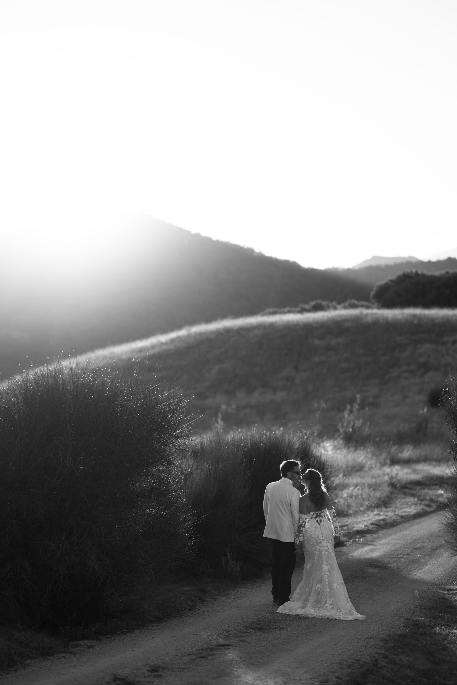 Black and white photo of a couple sharing a romantic moment on a dirt path surrounded by rolling hills at sunset.