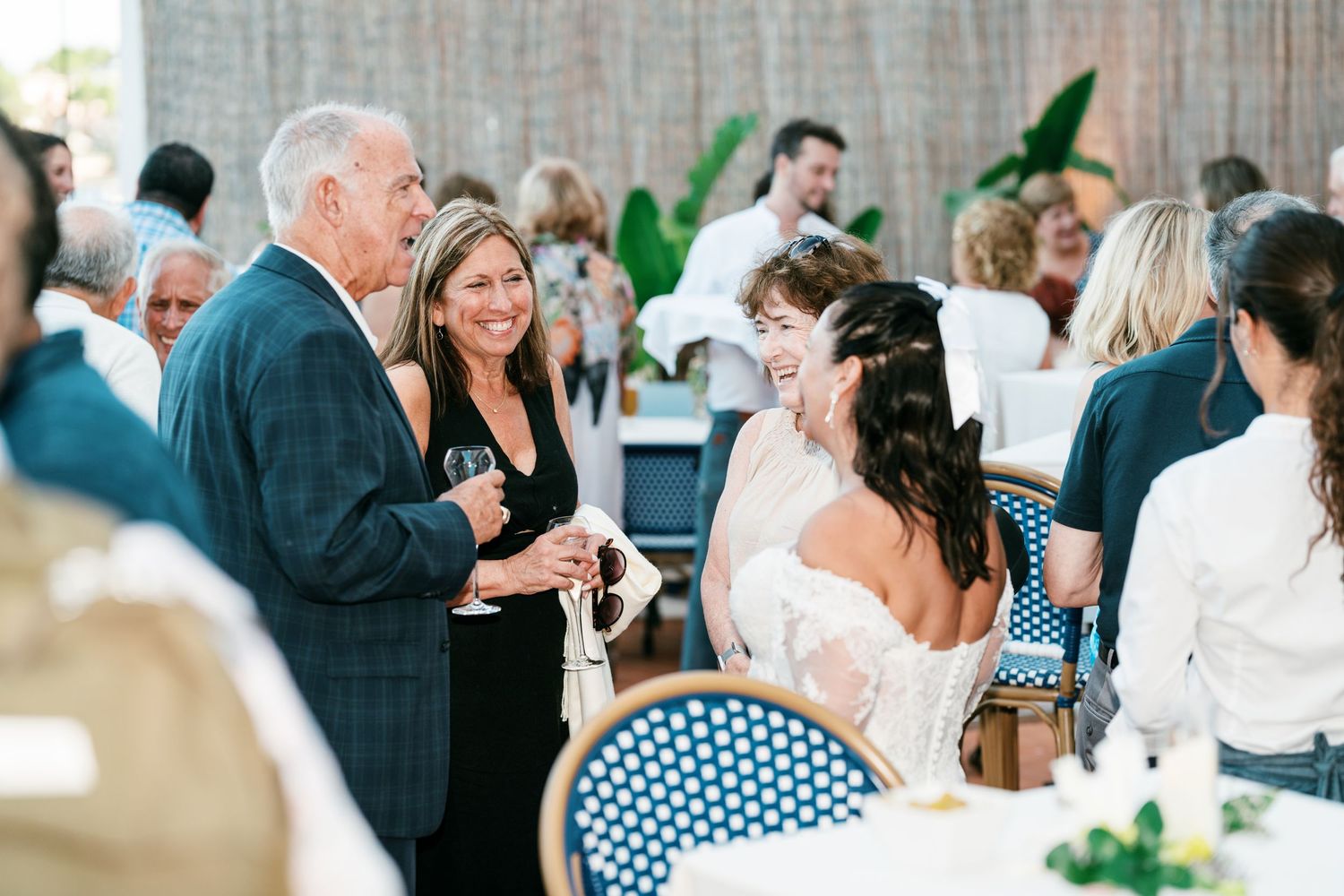 Smiling wedding guests mingle and chat with drinks in hand at a bright, festive reception with blue woven chairs and fresh greenery.