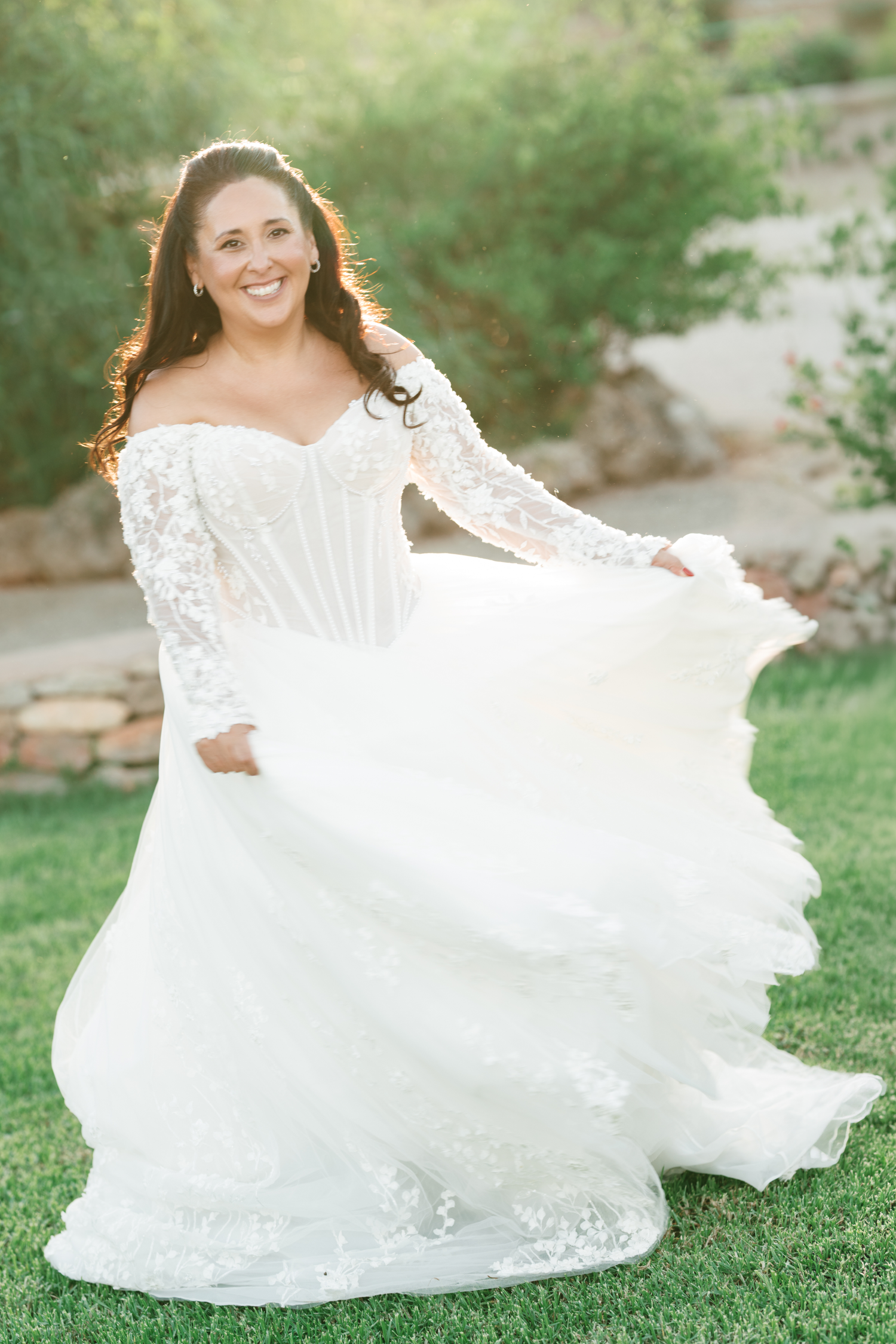 Smiling bride in an off-the-shoulder white lace wedding gown with a corseted bodice twirls her flowing tulle skirt on a sunny green lawn.