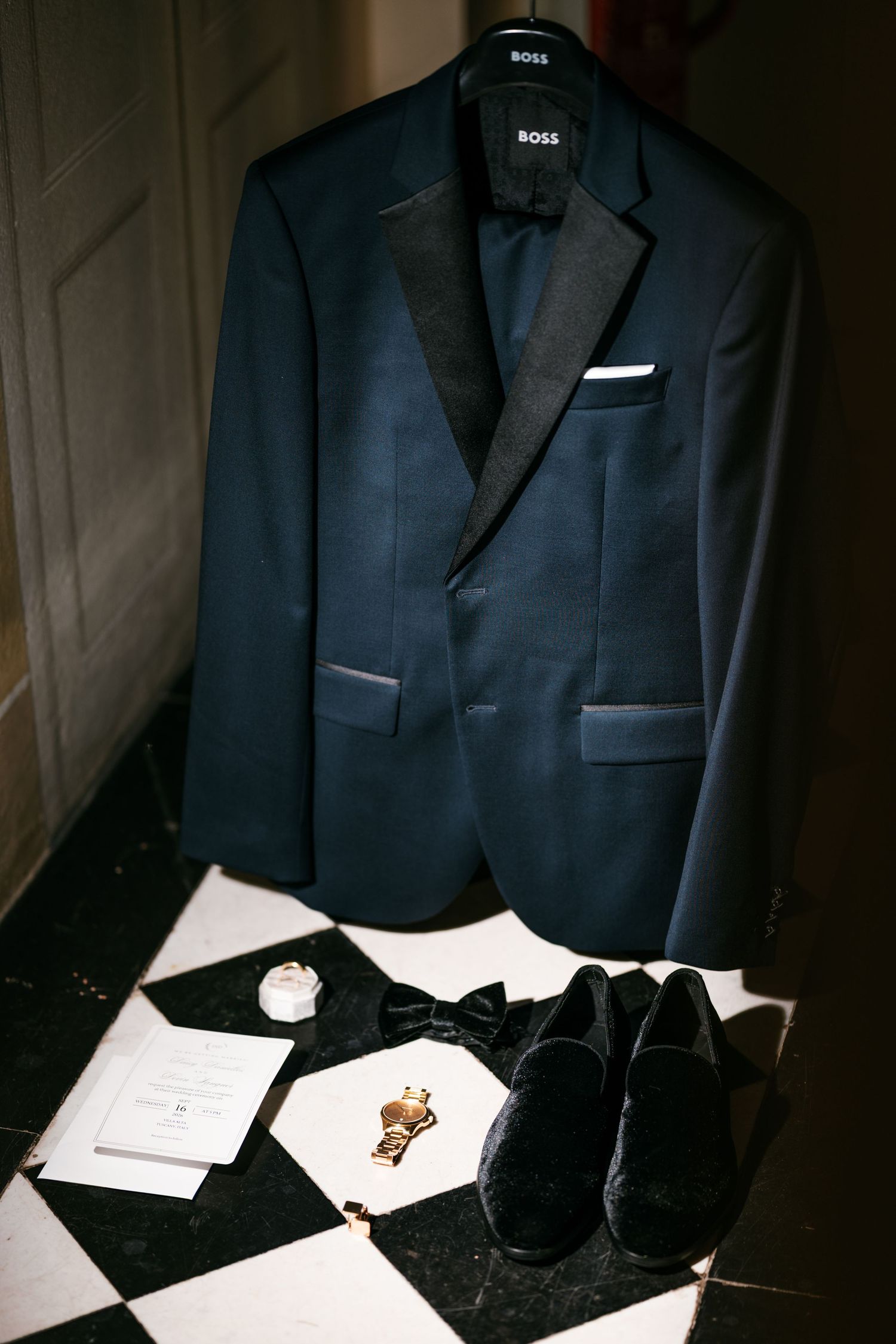 Navy Boss tuxedo jacket with black satin lapels hanging above a bow tie, velvet loafers, cufflinks and invitation on a checkered floor.