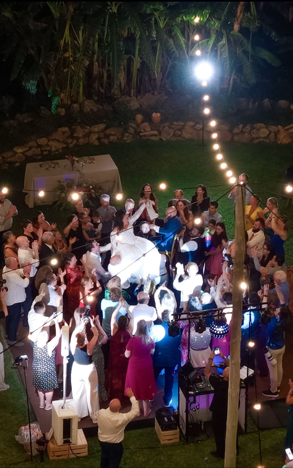 Wedding guests gather around a bride and groom dancing outdoors on a lawn at night, surrounded by string lights and tropical foliage.
