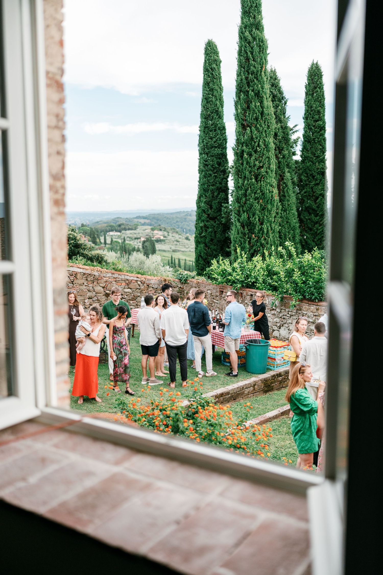 Wedding guests mingle at an outdoor garden reception, viewed through a window, with tall cypress trees and Tuscan hills in the background.