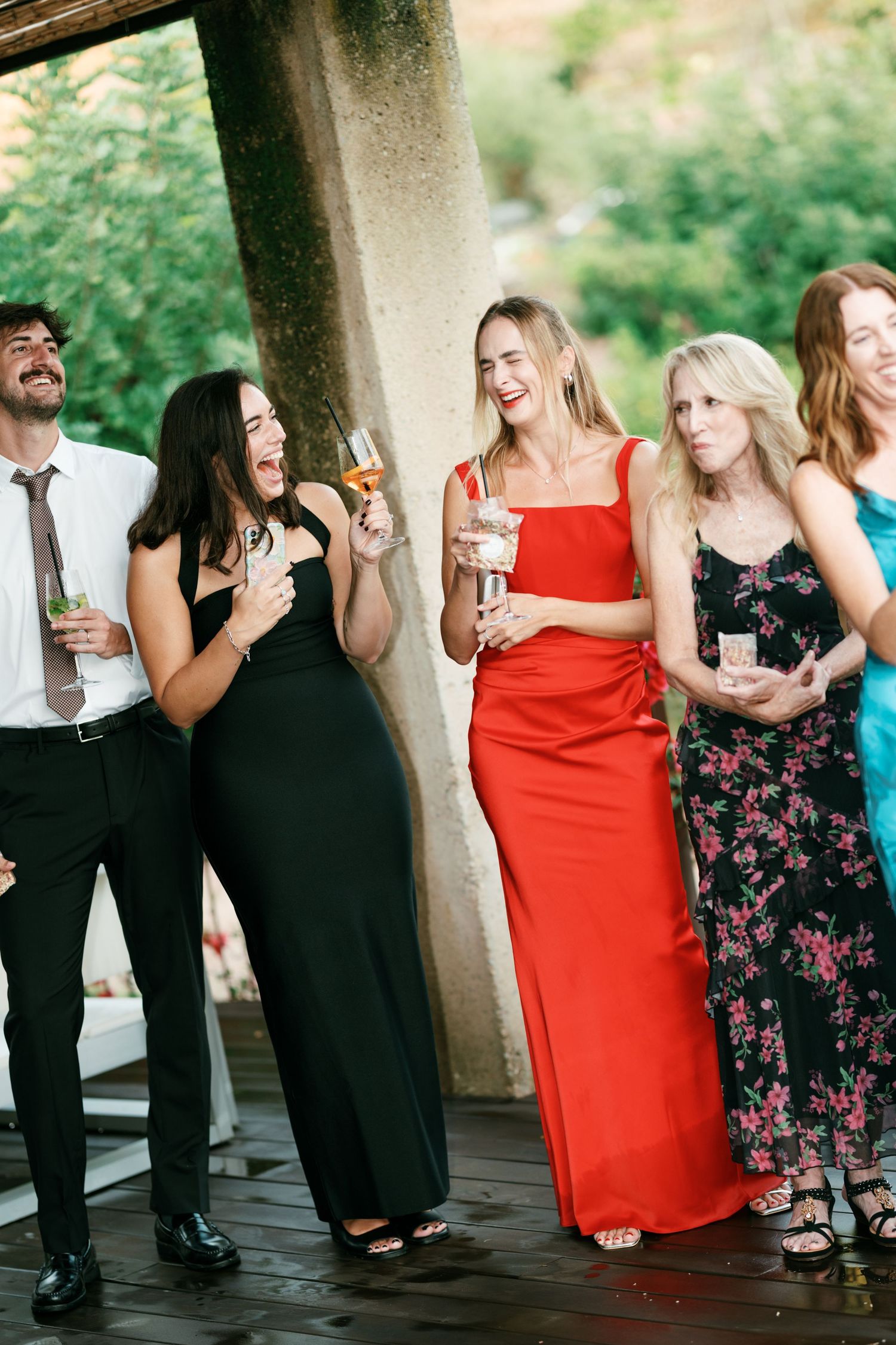Five guests in elegant formal attire laugh and hold drinks while celebrating together on a wooden deck at an outdoor wedding reception.