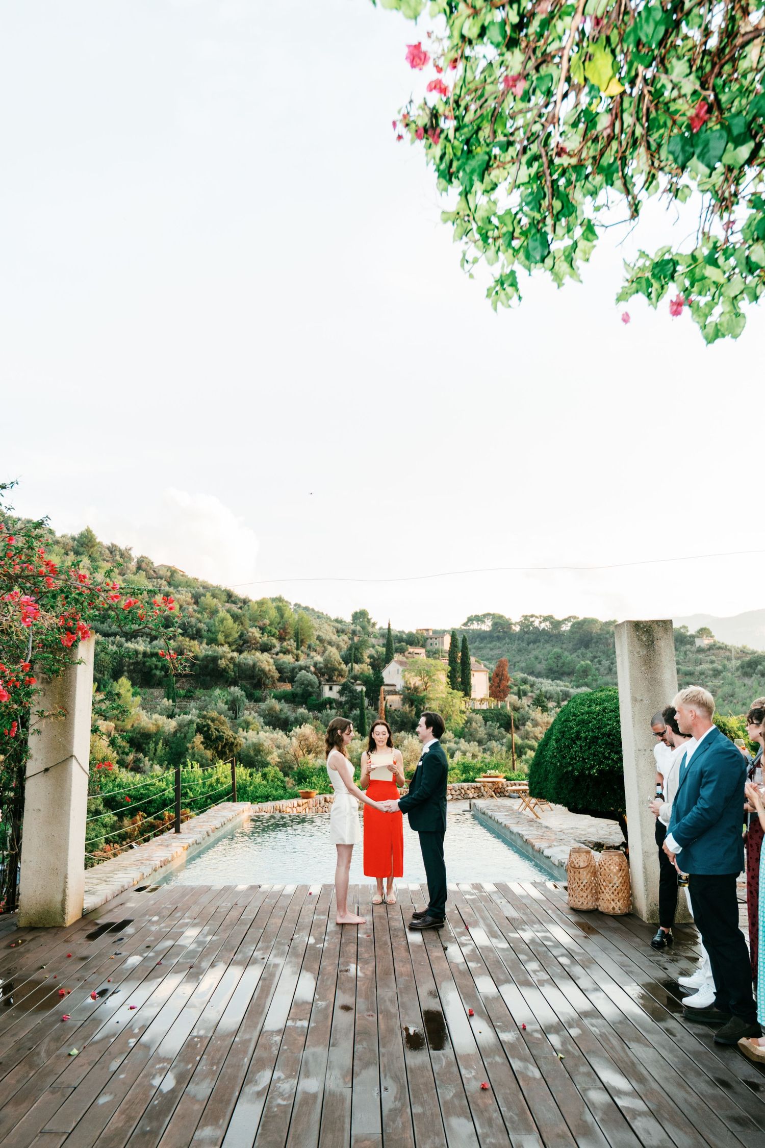 Couple exchanges vows with an officiant on a wet wooden deck beside a pool, with guests and lush green hills in the background.