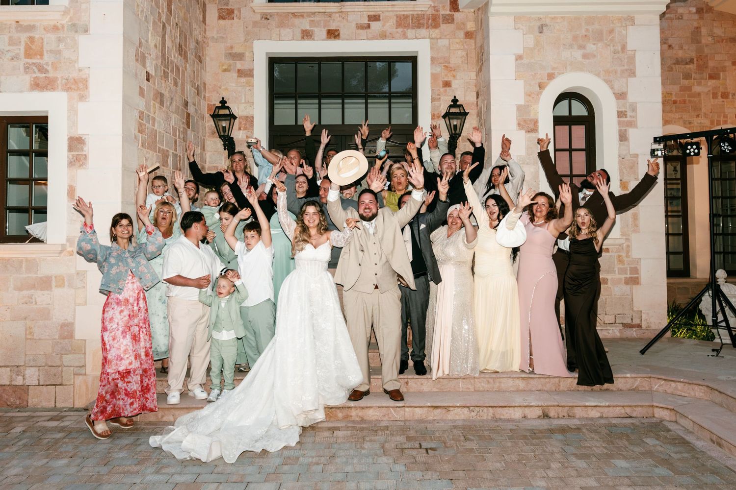 Wedding party celebrates on stone steps with bride and groom, all raising arms joyfully at outdoor venue.