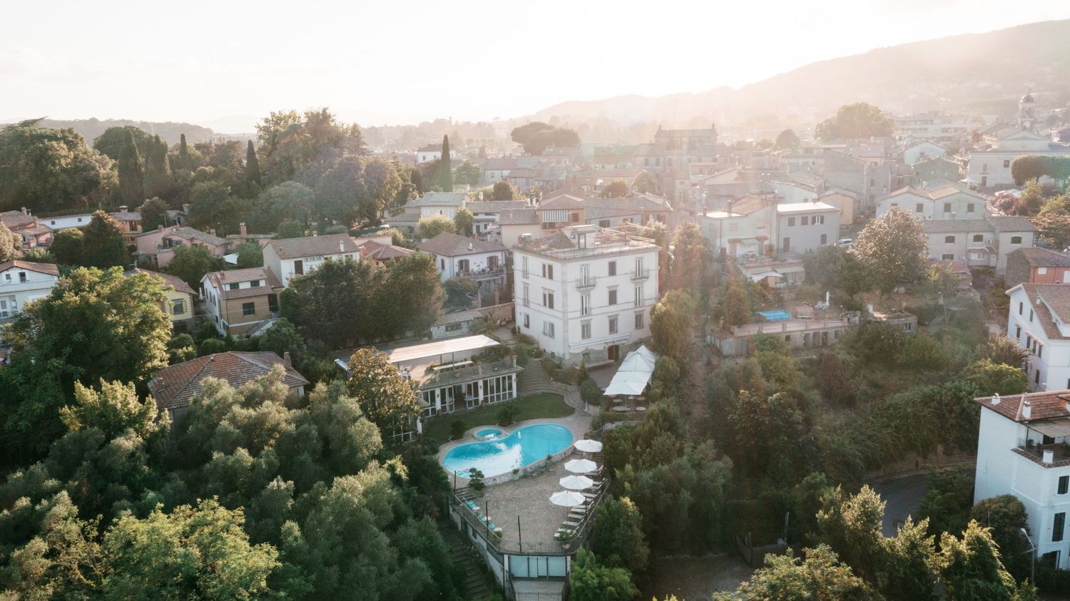 Aerial view of a Mediterranean villa with a swimming pool surrounded by lush trees and buildings at golden hour. Tuscany wedding photograp