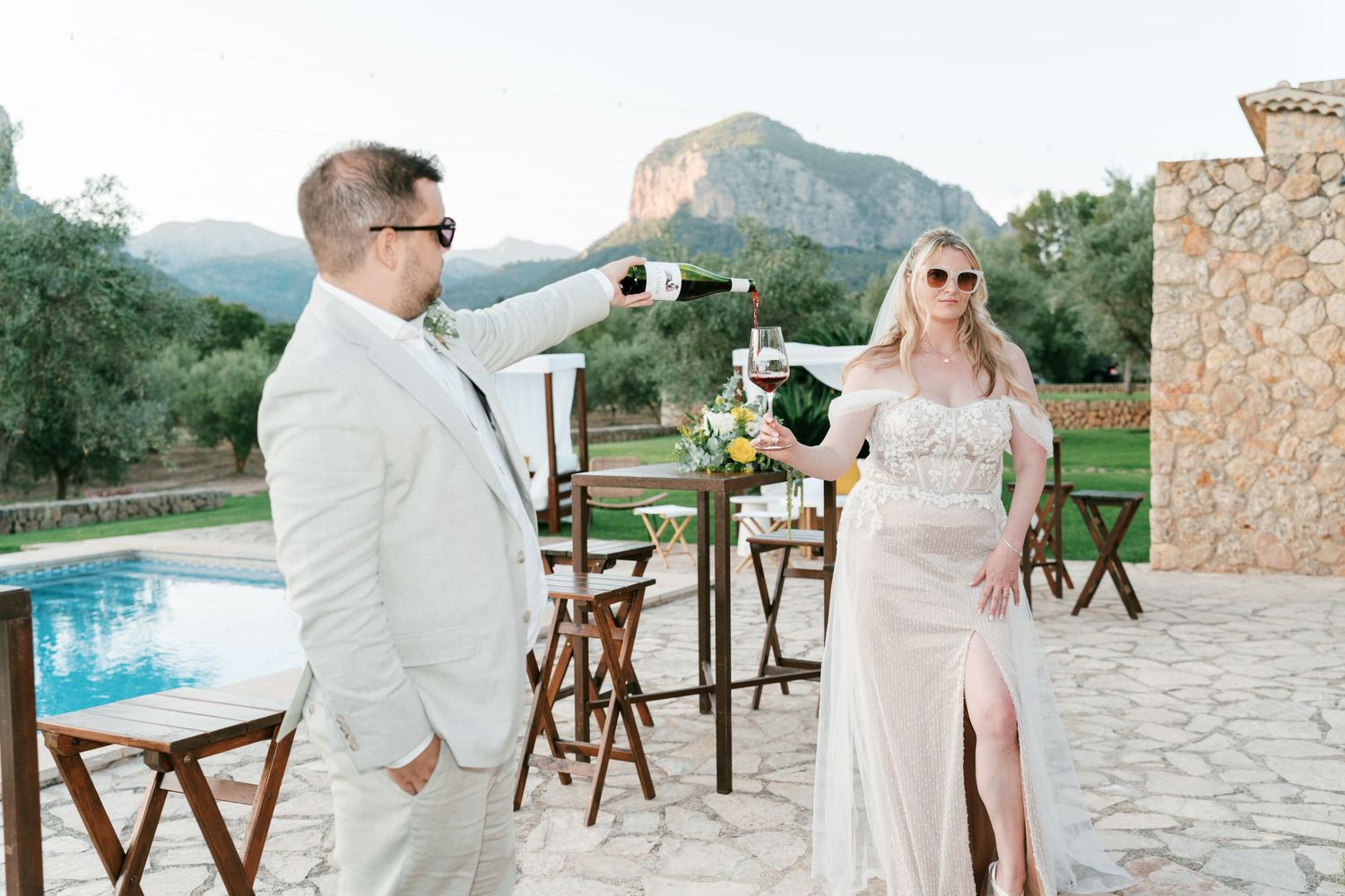 Couple celebrating outdoors by pool with champagne, mountains in background, groom in white suit, bride in fitted gown. Mallorca wedding.