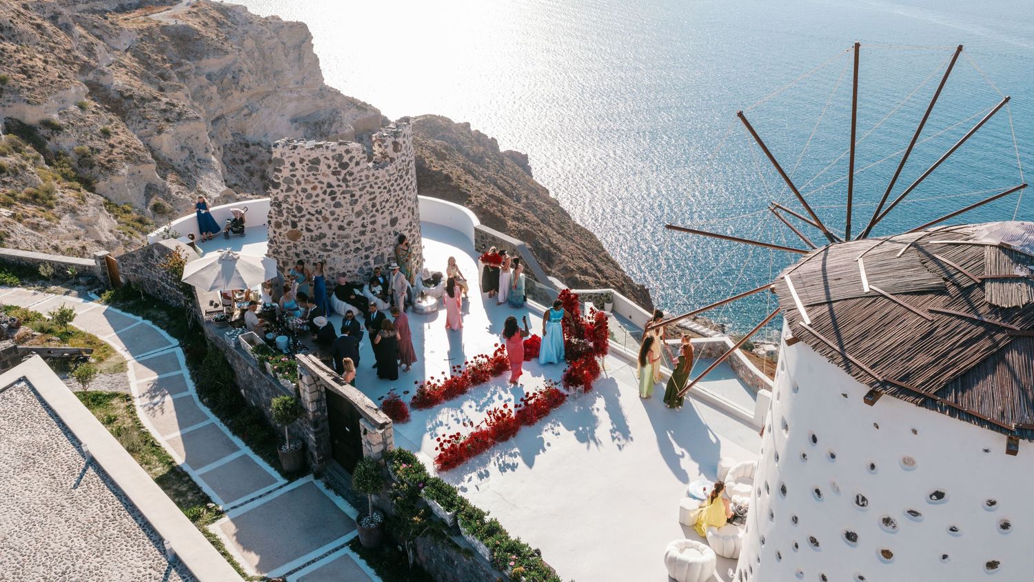 Aerial view of a wedding ceremony at a scenic cliffside windmill venue in Santorini, Greece, overlooking the blue Aegean Sea.