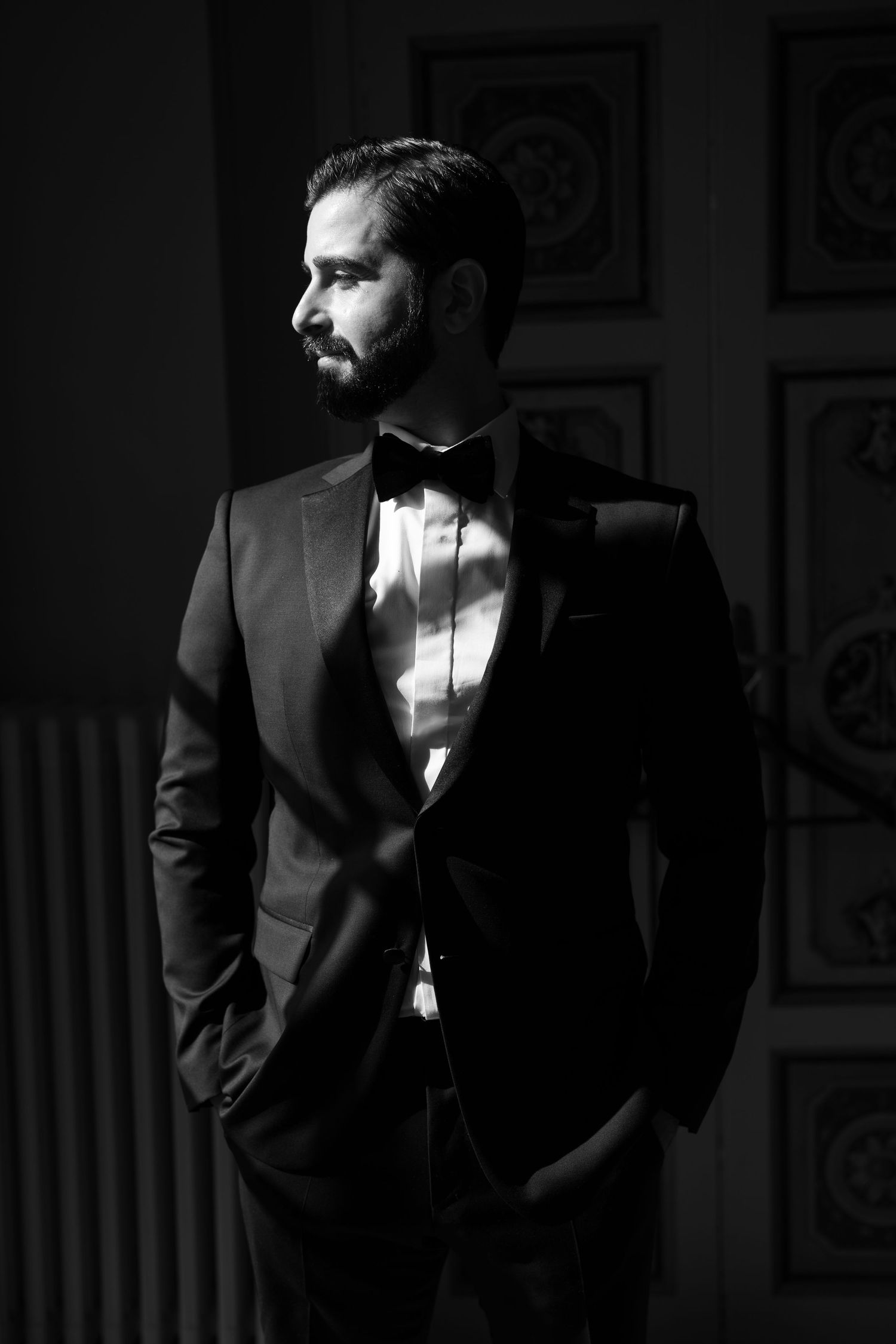 Black and white portrait of a bearded man in a tuxedo and bow tie standing in dramatic window light with hands in his pockets.