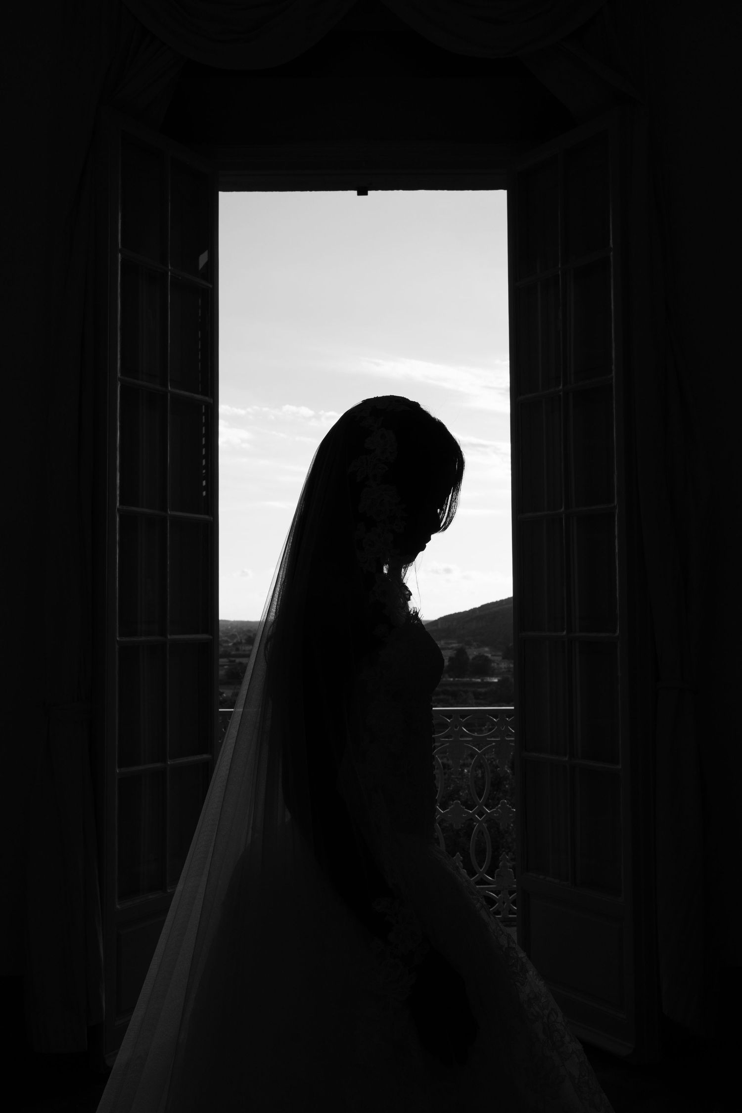 Black and white silhouette of a bride in a long veil standing before open balcony doors with a scenic hillside view.
