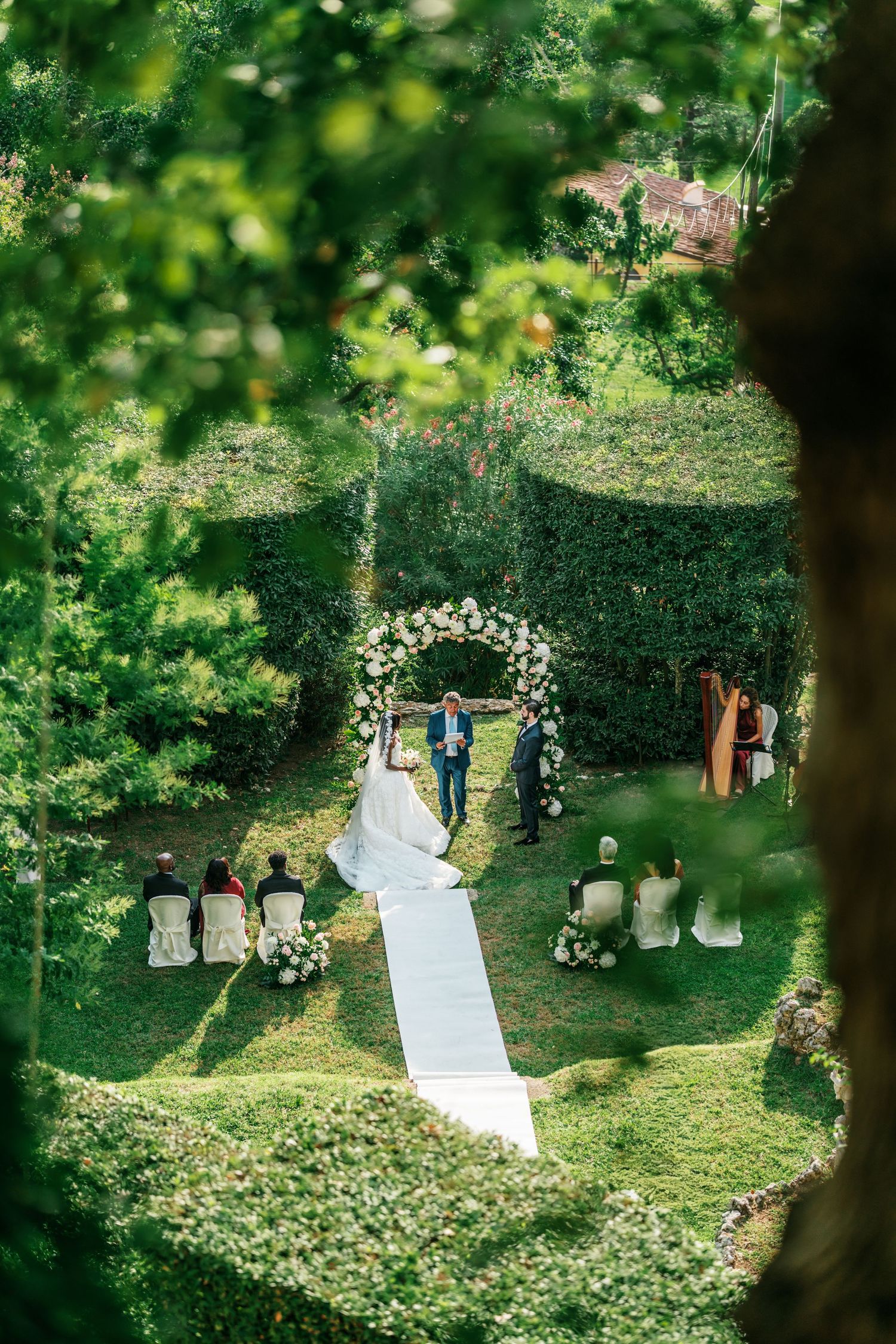 Outdoor garden wedding ceremony with a bride in a long gown at a floral arch, guests seated on white chairs along an aisle.