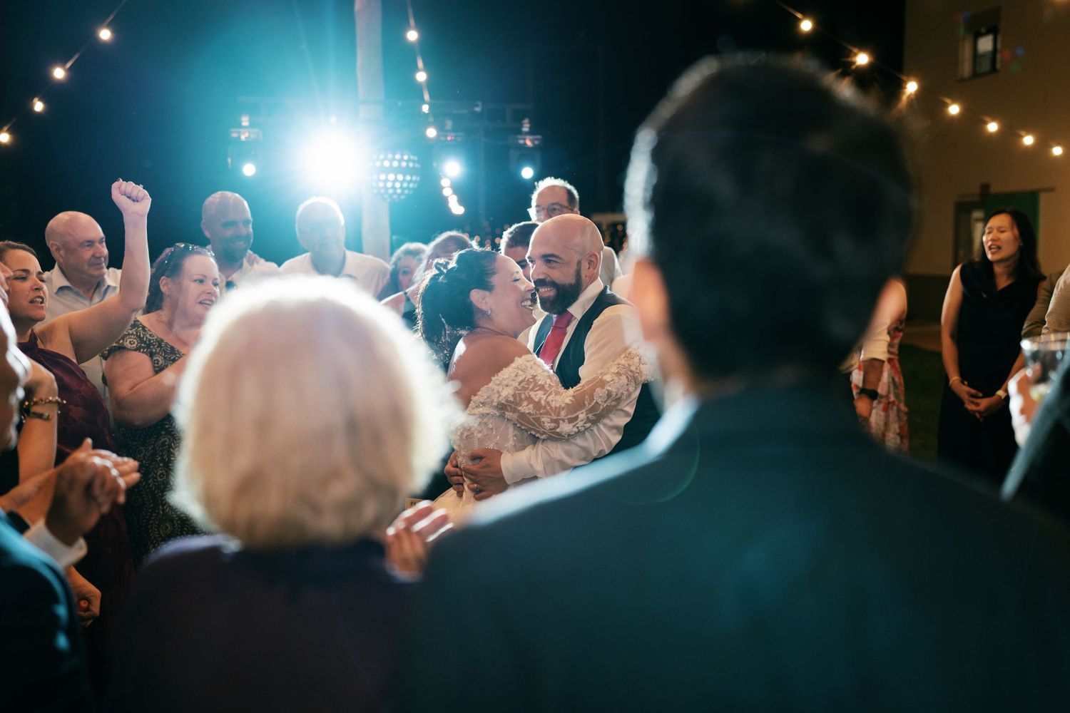 Bride and groom share a first dance at an evening wedding reception as smiling guests watch beneath warm string lights.