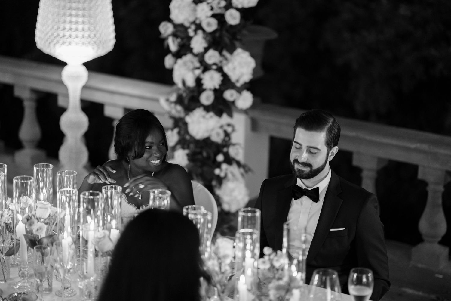 Smiling woman and bearded man in a tuxedo and bow tie sit at an elegant wedding reception table with white flowers and crystal glassware.
