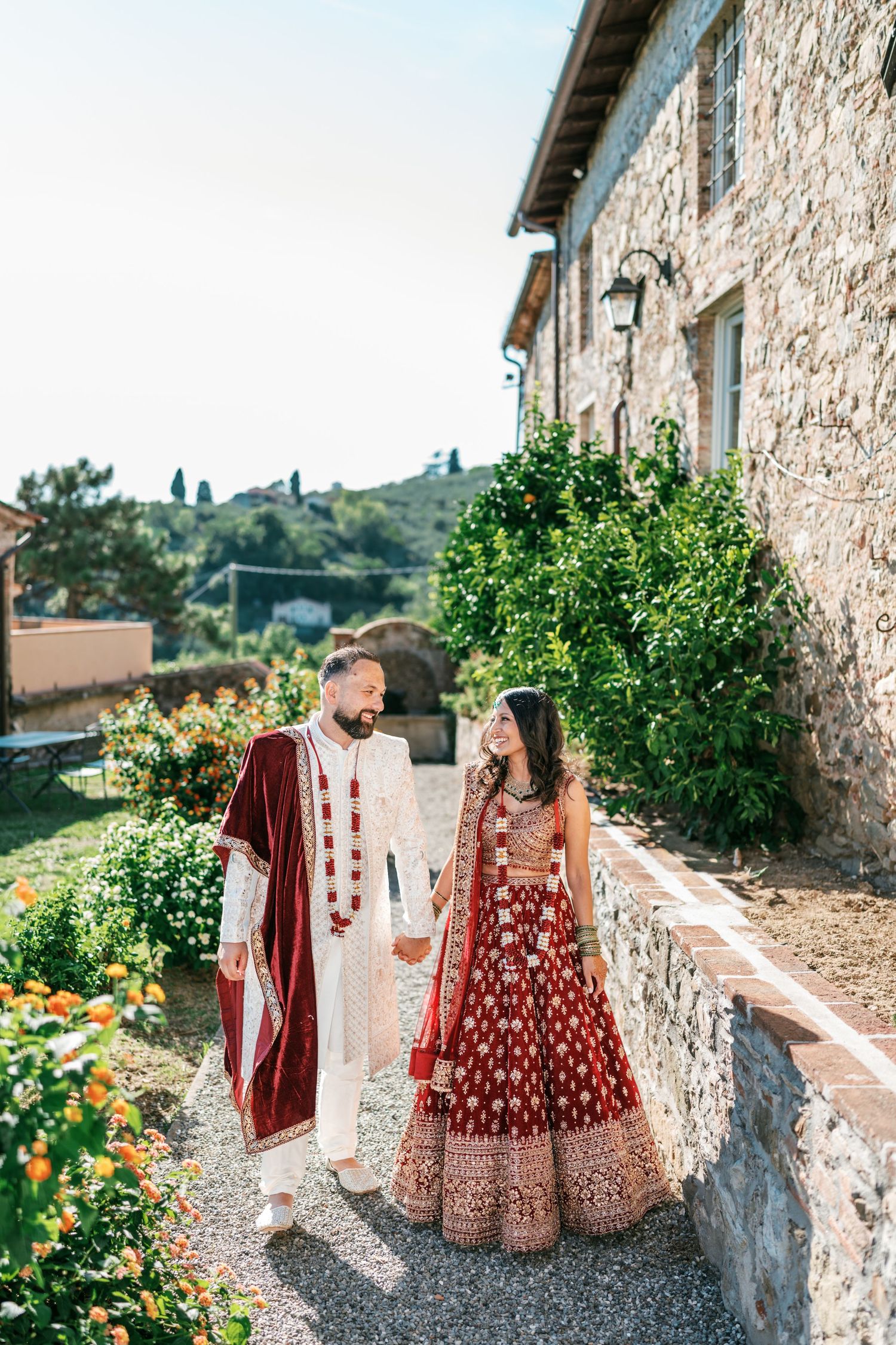 Indian bride and groom in red and ivory wedding attire holding hands on a gravel path beside a rustic stone villa in Tuscany.