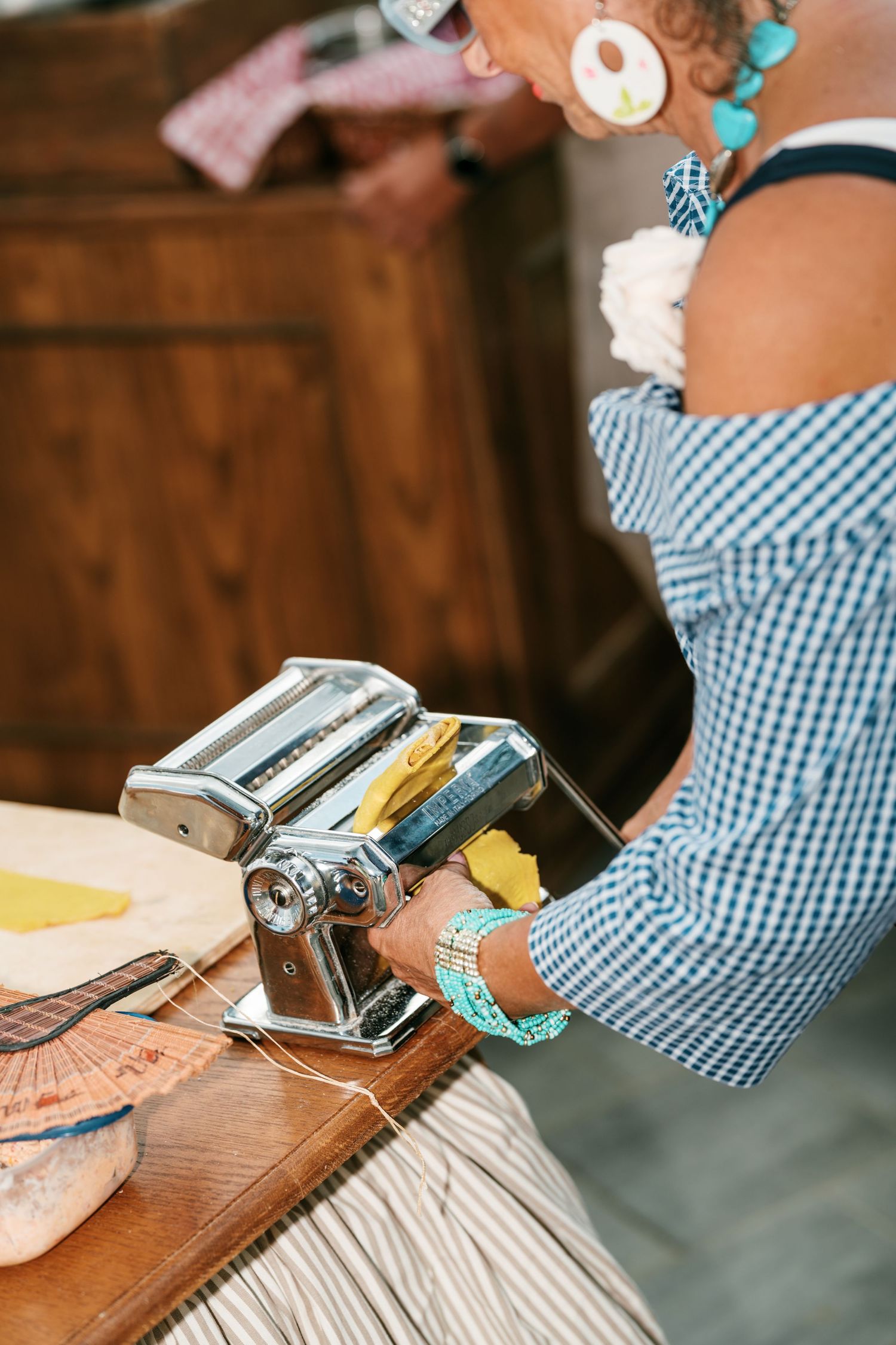 Woman in a gingham off-shoulder top and turquoise jewelry rolls fresh yellow pasta dough through a stainless steel pasta machine.