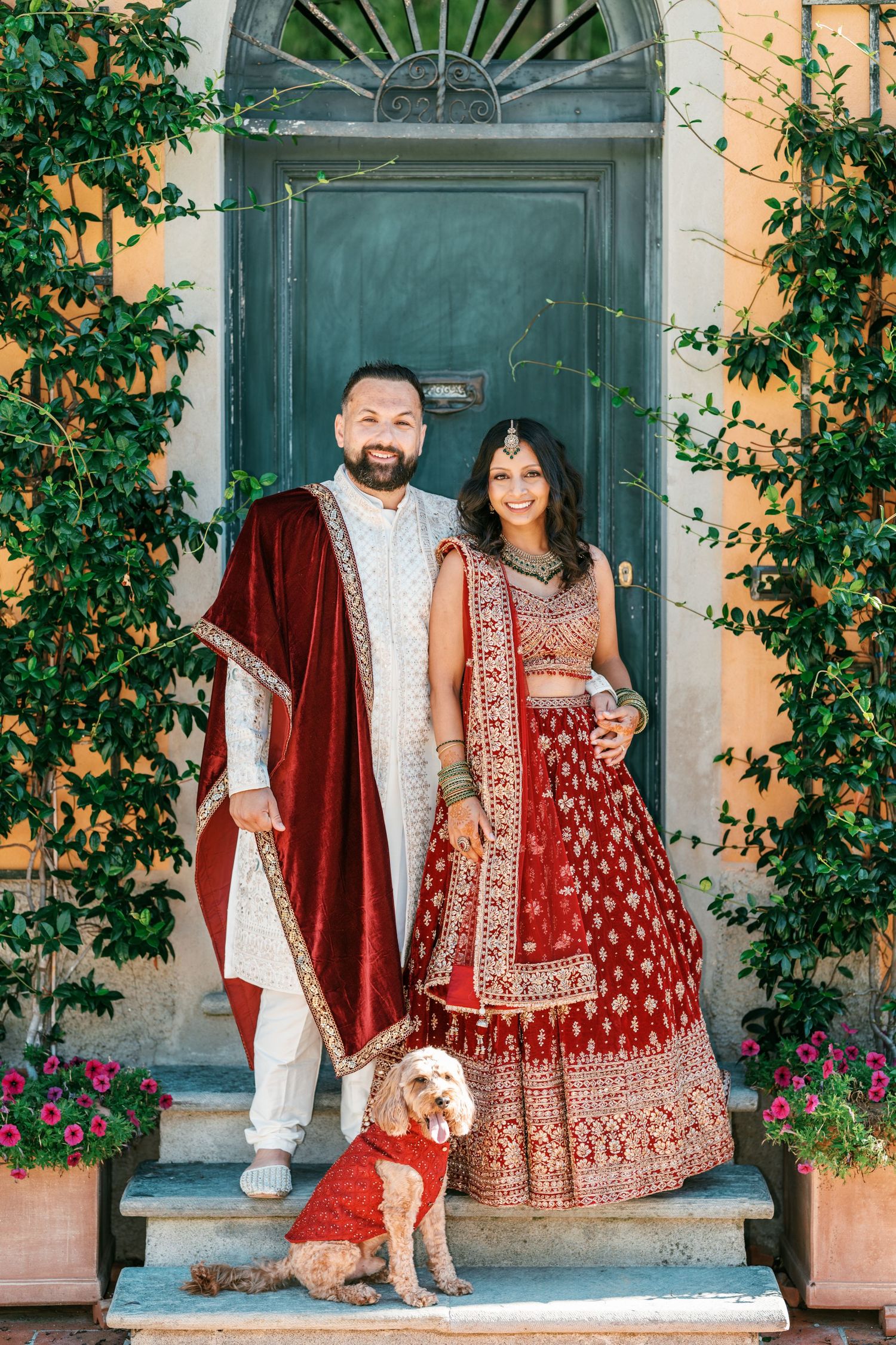 Couple in red and gold traditional Indian wedding attire pose with their golden doodle dog in matching outfit on steps before a green door.
