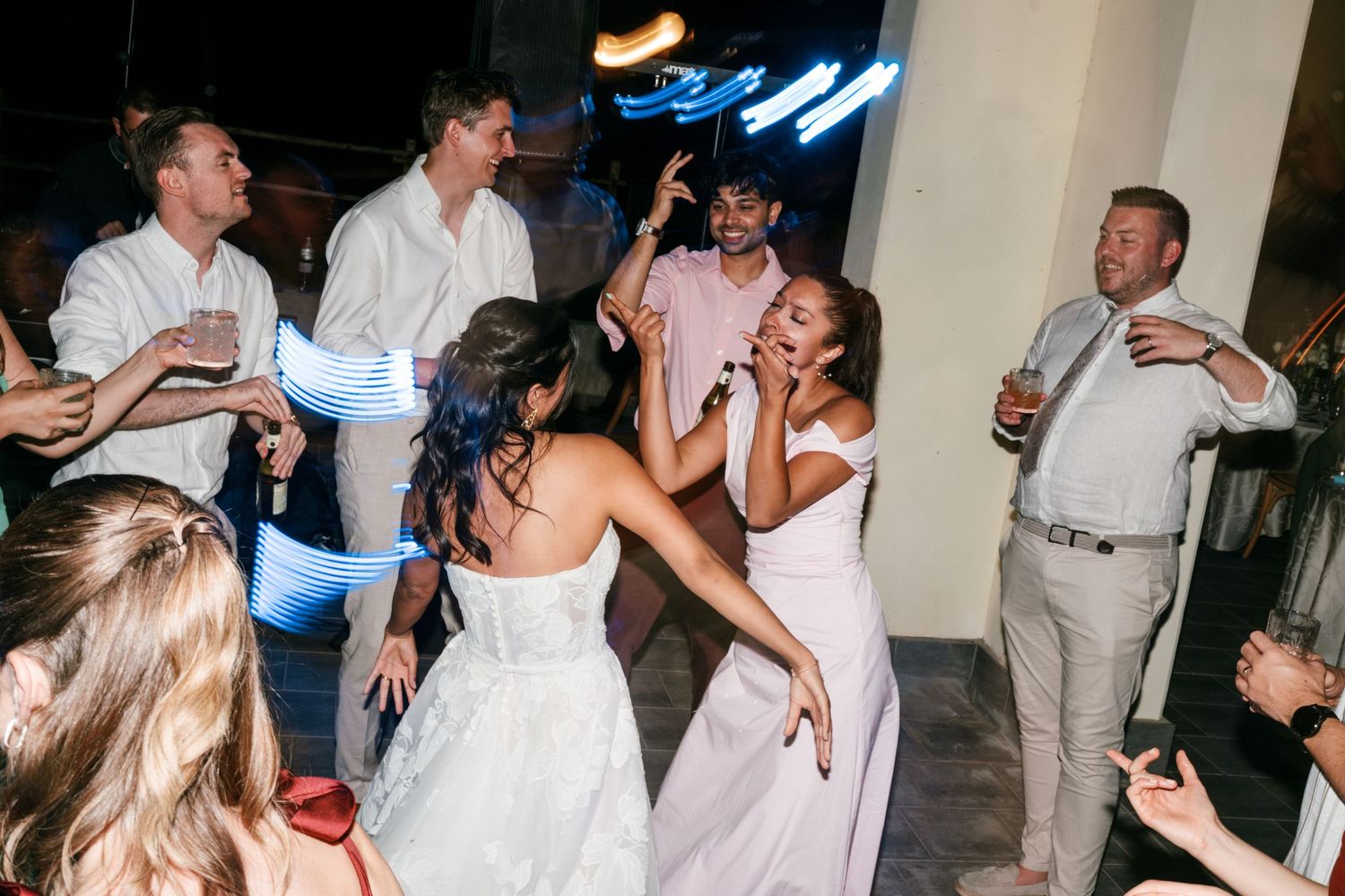 Wedding guests in light summer attire dance and celebrate with a bride in a white gown on a lively dance floor under colorful party lights.